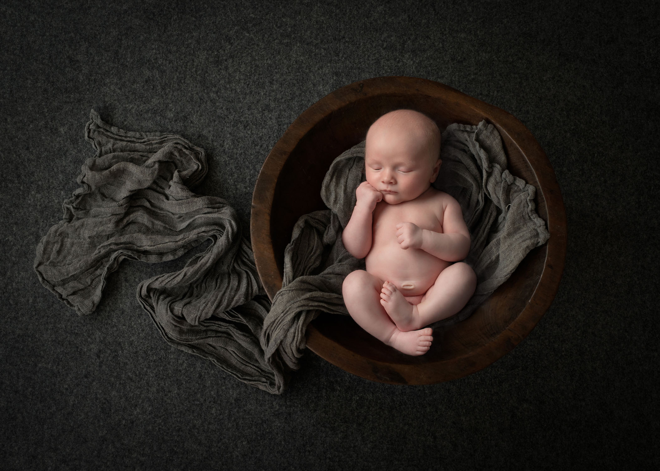 newborn boy sleeping in wooden bowl with grey wrap laying around him
