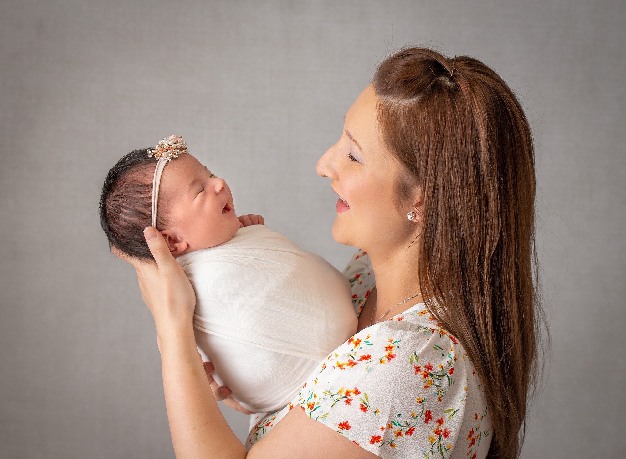 mother smiling at newborn baby girl swaddled in white wearing floral headband on gray backdrop