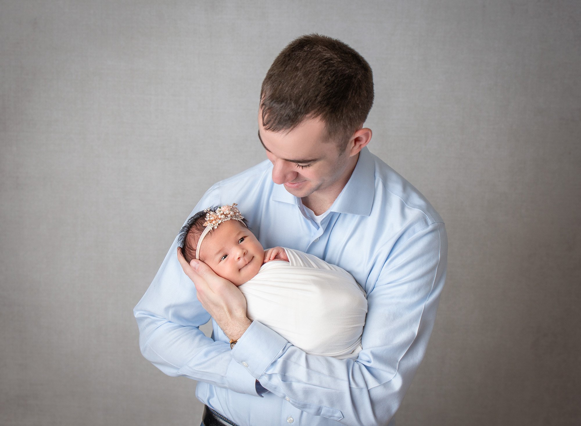dad in light blue button up looking down at his newborn baby girl swaddled in white wearing floral headband