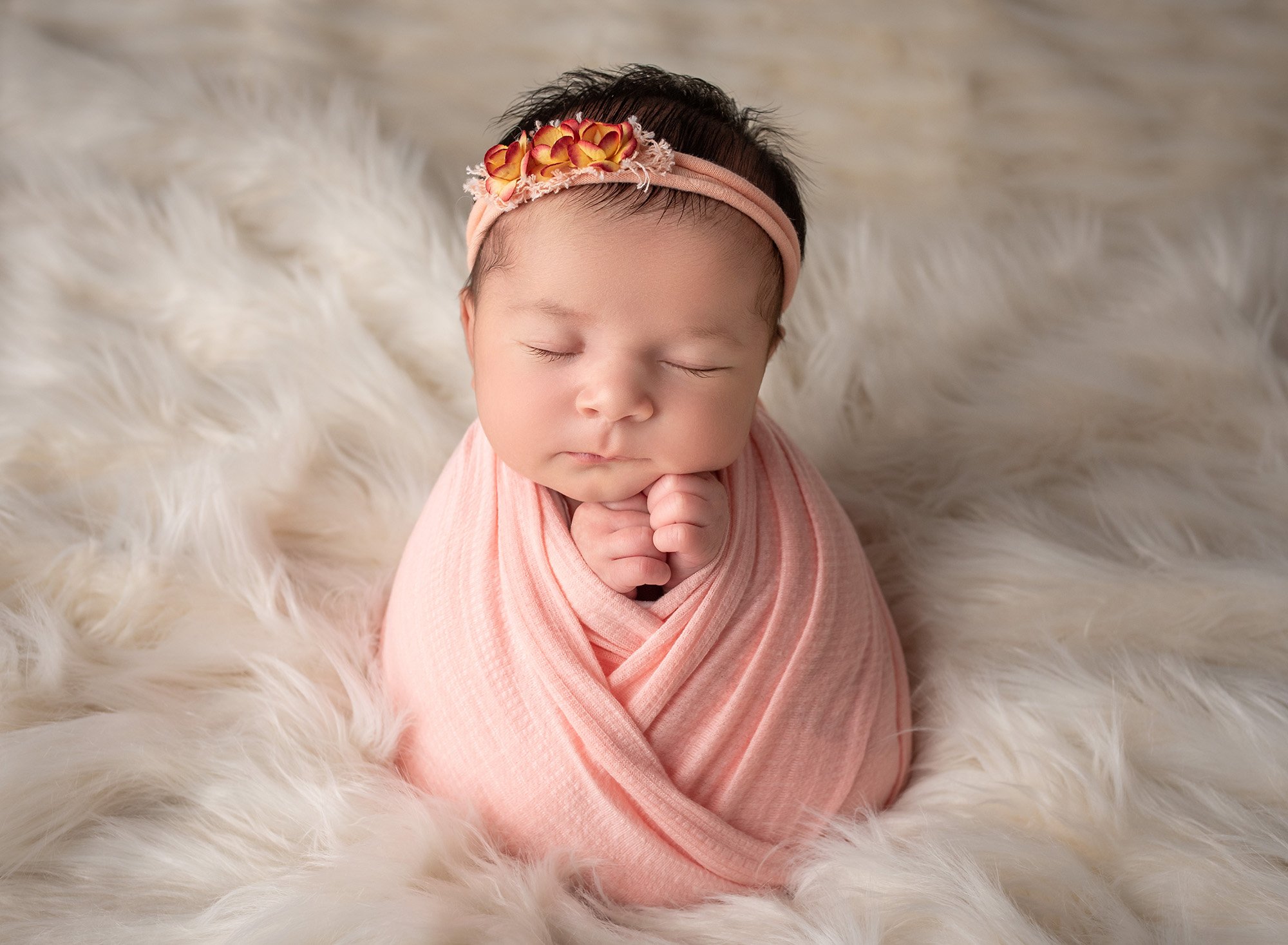 brunette baby girl swaddled in peach with headband on fluffy cream blanket