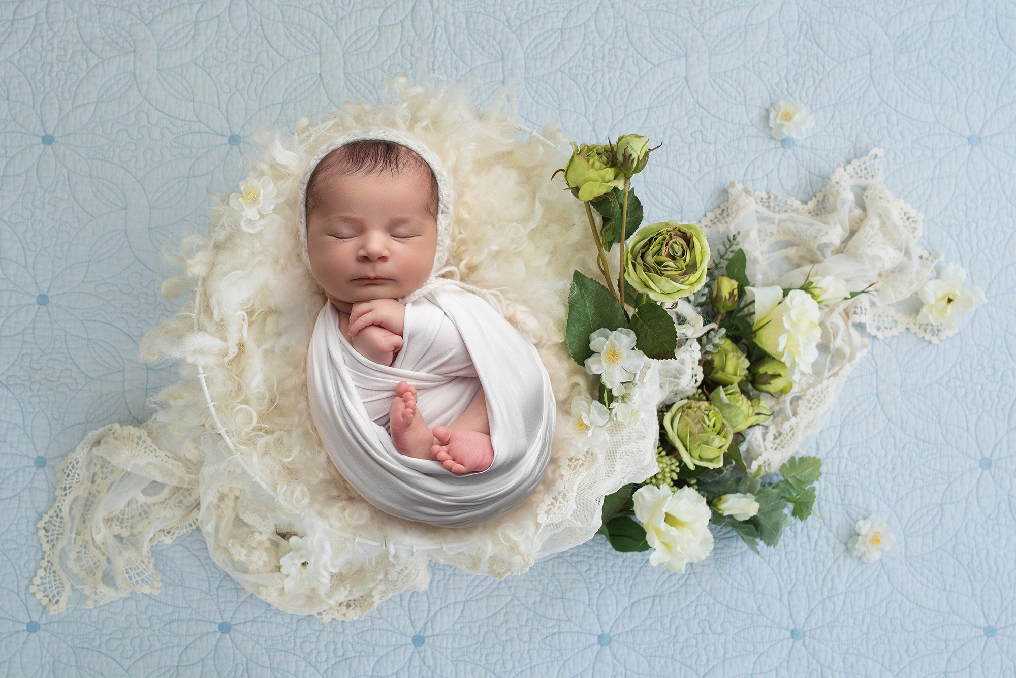 baby girl swaddled in white laying on top of antique lace blanket with white and green floral accents on floral background
