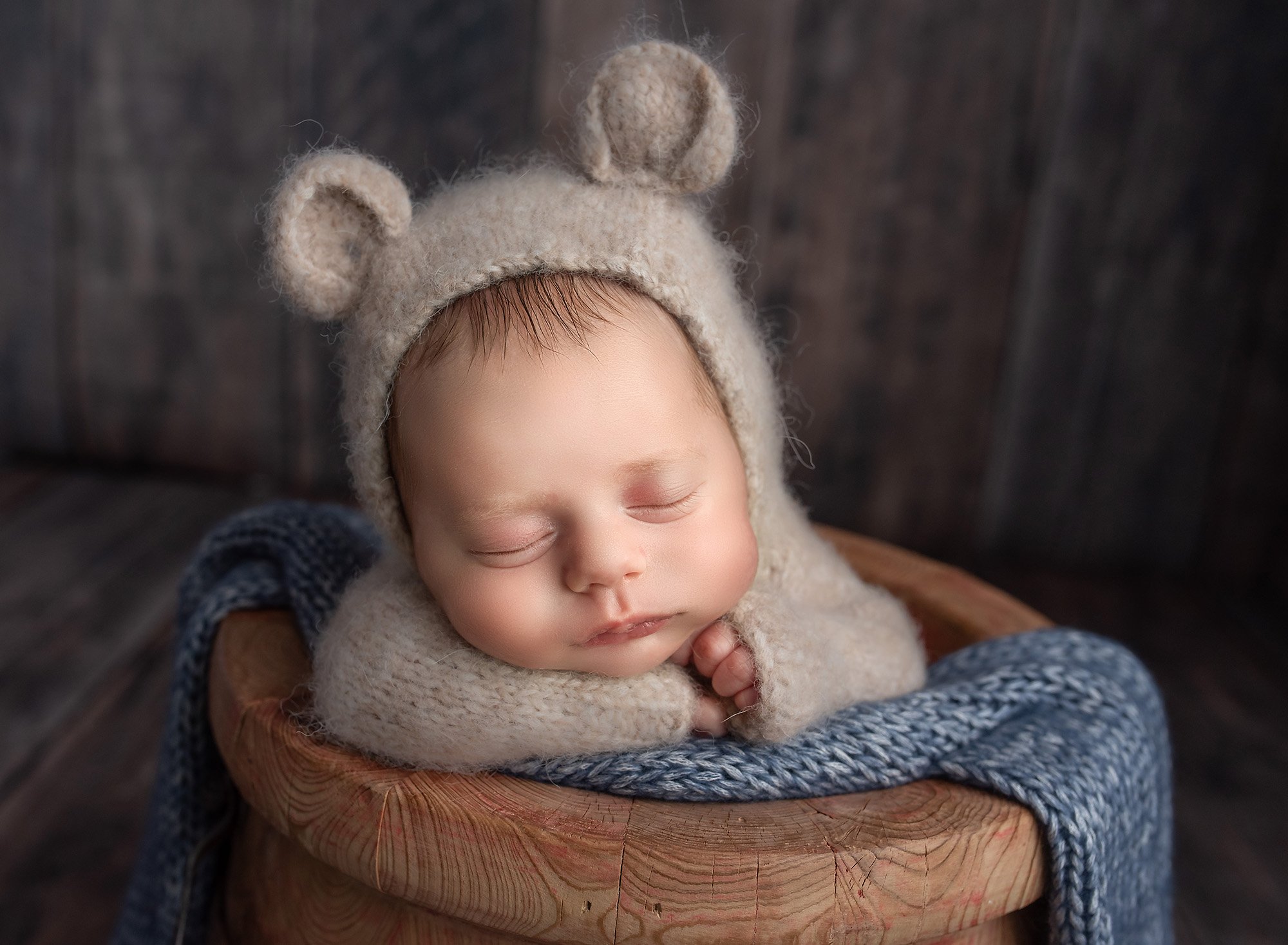 award winning newborn photography newborn baby boy dressed in bear sweater romper sitting in wooden bowl with blue sweater blanket