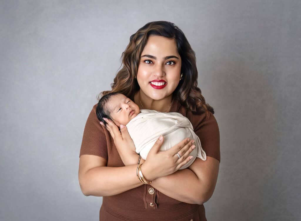 newborn and family photography portrait of mother holding swaddled baby against gray backdrop