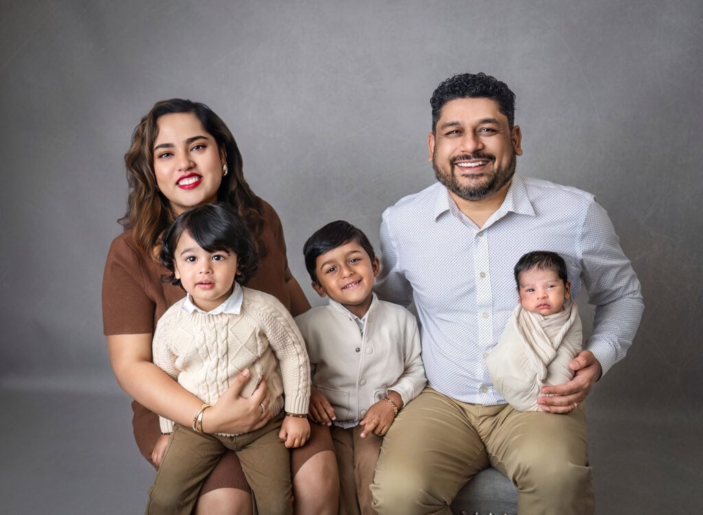 newborn and family photography with parents, two young boys, and swaddled baby on neutral backdrop