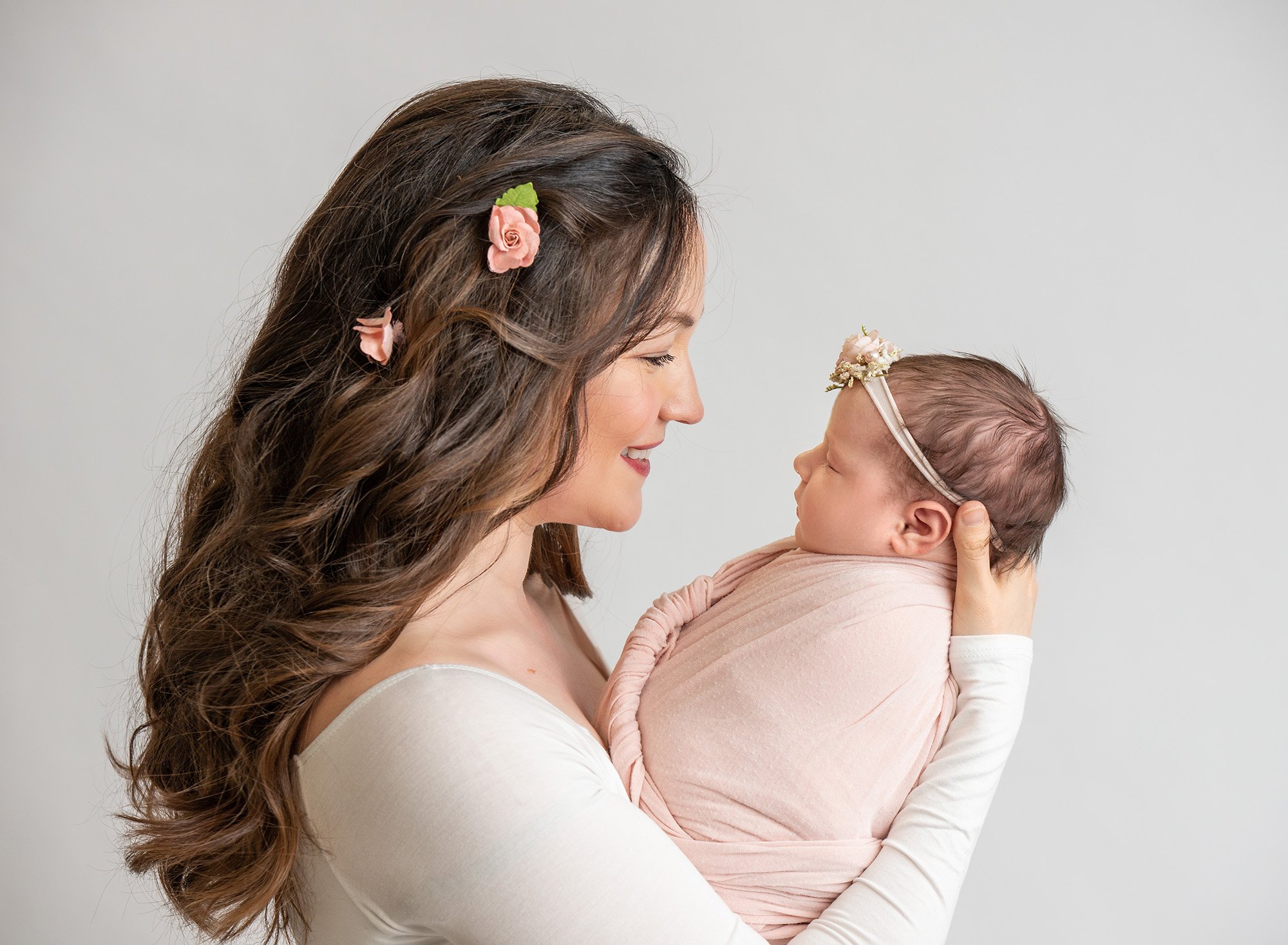 brunette with pink flowers in hair looking at newborn baby girl swaddled in soft pink