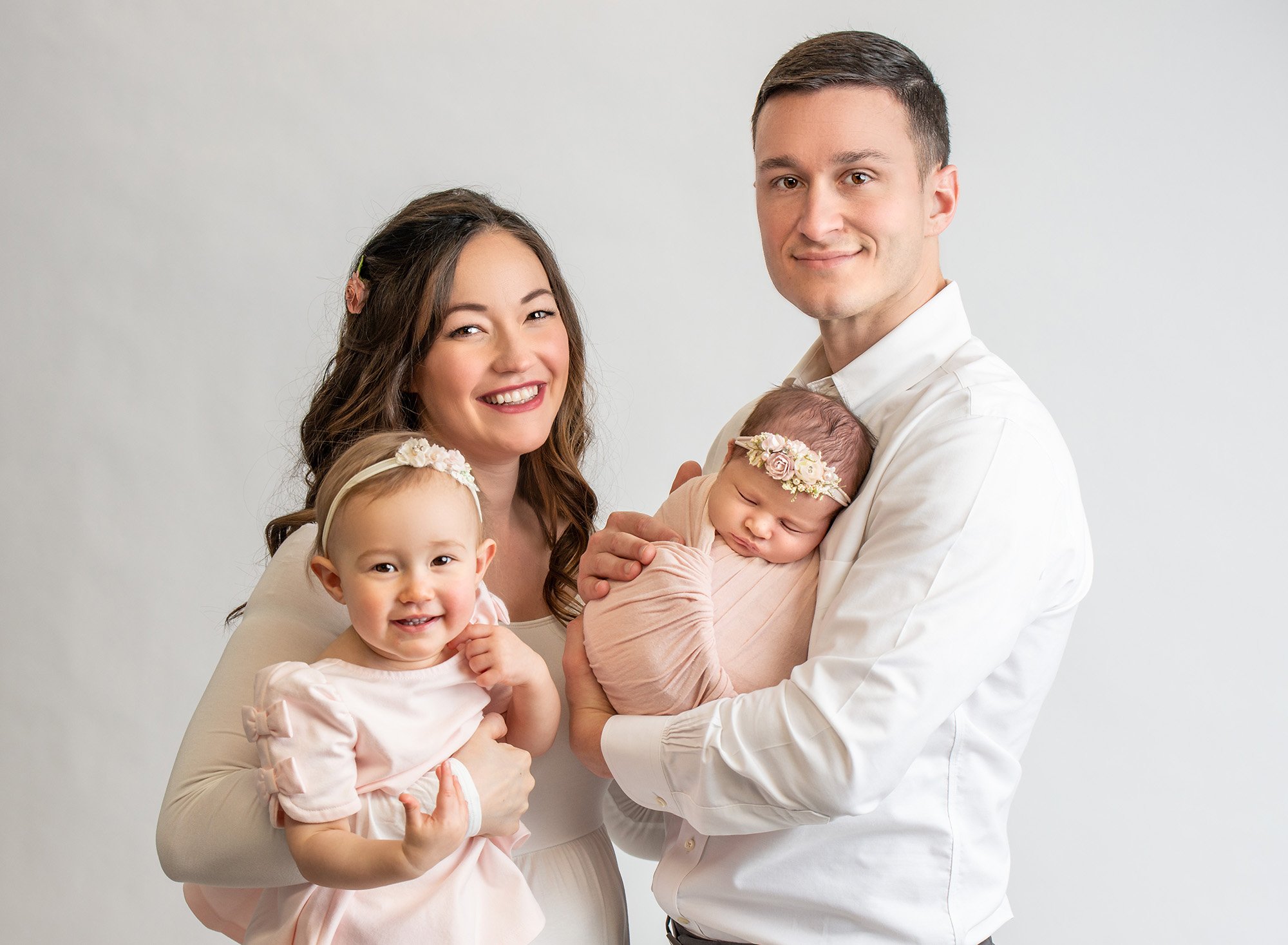 mom and dad on white backdrop holding young daughter and newborn daughter in pink