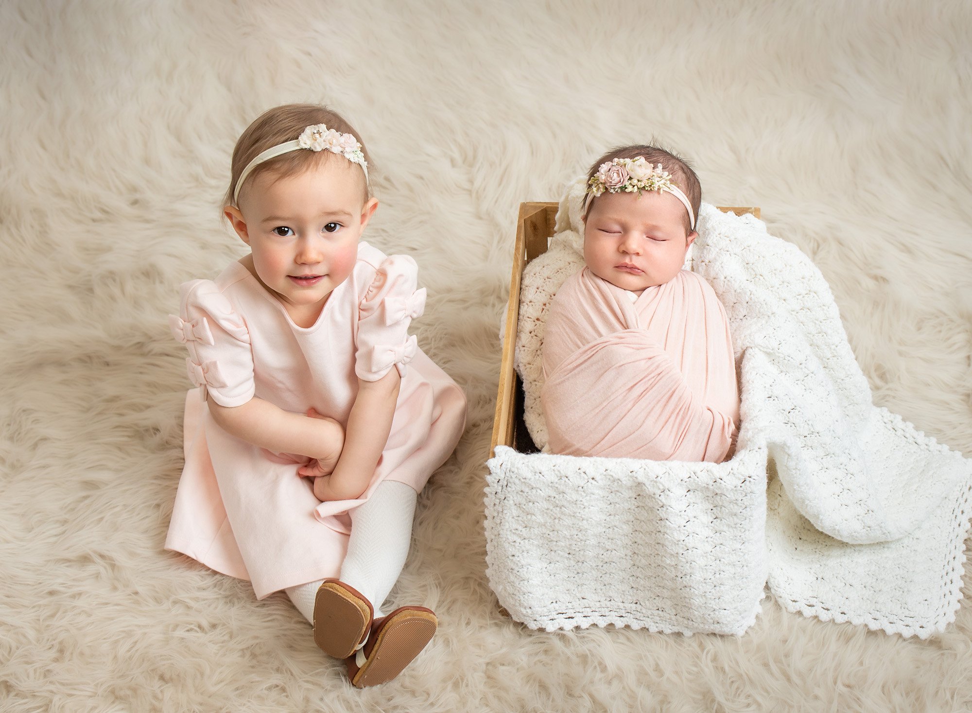 newborn baby swaddled in pink and floral headband laying in wooden box with white blanket next to lovely big sister in pink dress on fluffy blanket