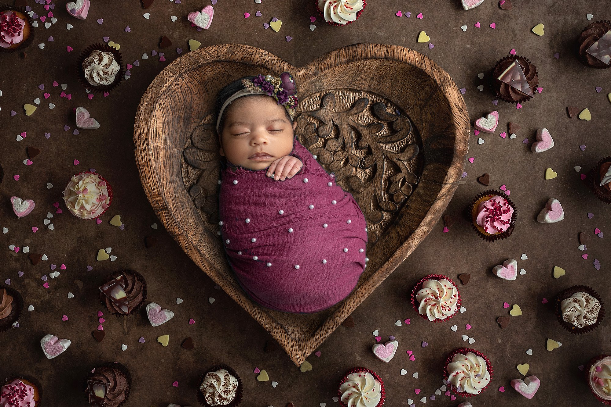 sleeping newborn baby girl swaddled in magenta wrap with pearls wearing purple floral headband laying in wooden heart bowl surrounded by cupcakes and heart candies