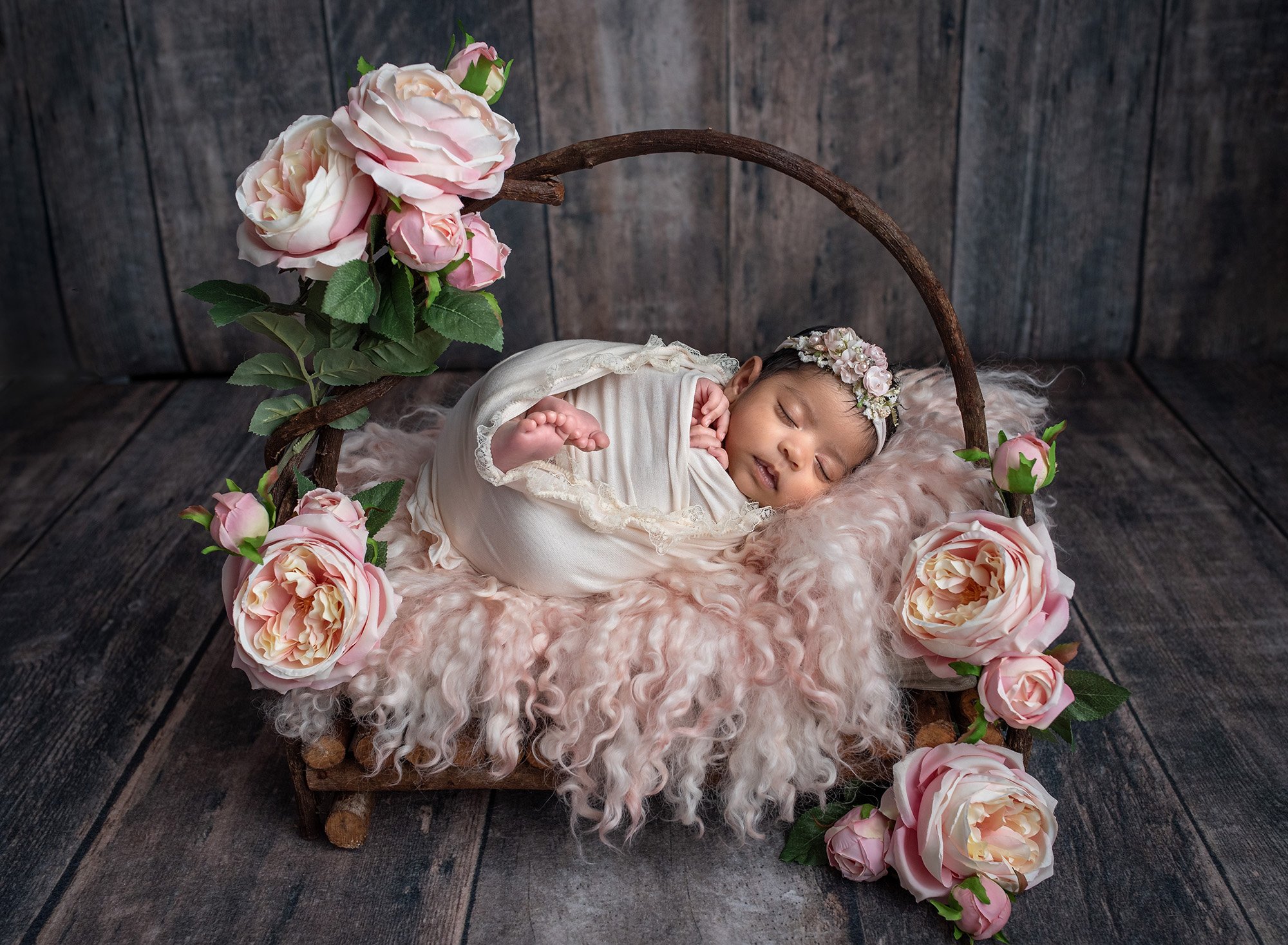 newborn baby girl swaddled in lace wrap sleeping on top of rustic wooden bed with pink fuzzy blanket surrounded by pink flowers on wooden background