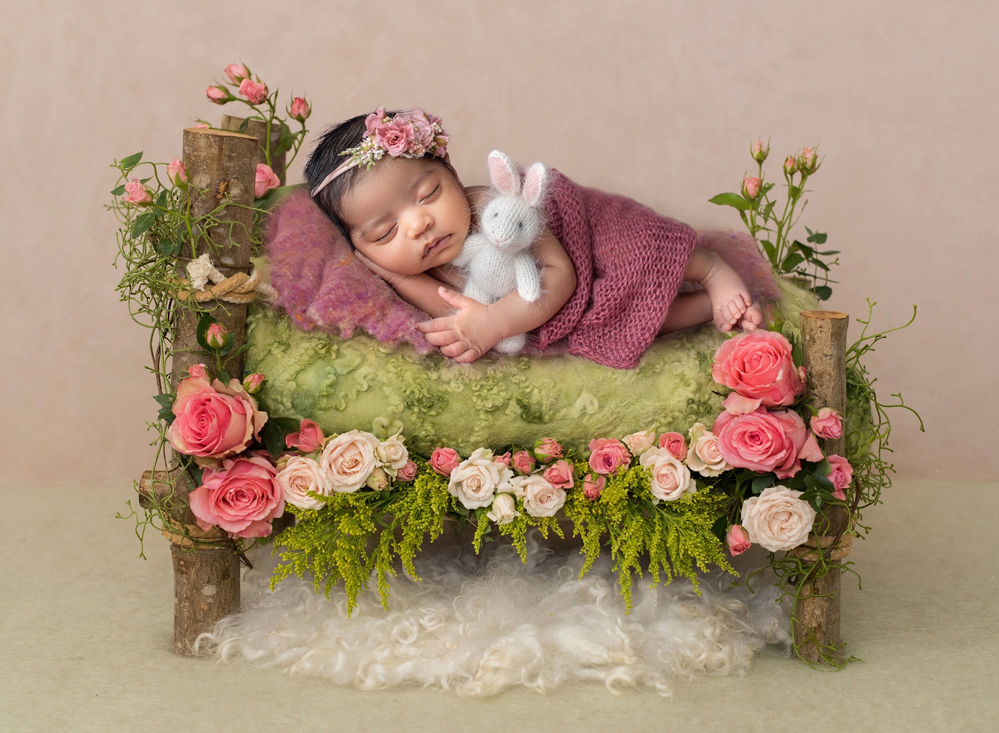 newborn baby girl sound asleep on wooden bed draped in mauve sweater wrap holding stuffed bunny surrounded by pink flowers and greenery atop a fuzzy blanket