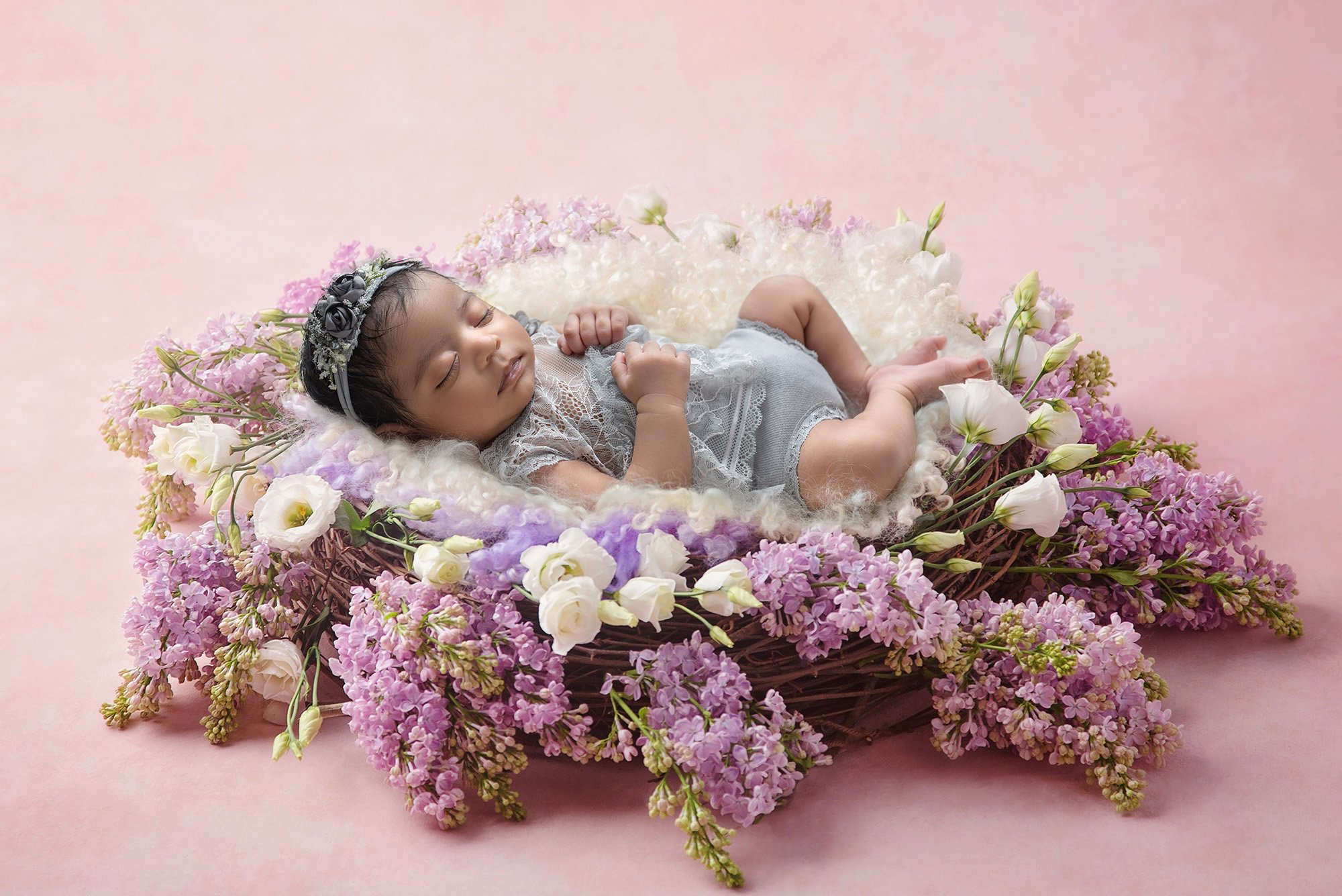 newborn baby girl in laced grey dress sound asleep on top of a purple and white floral wreath with fuzzy purple and white blankets on pink background