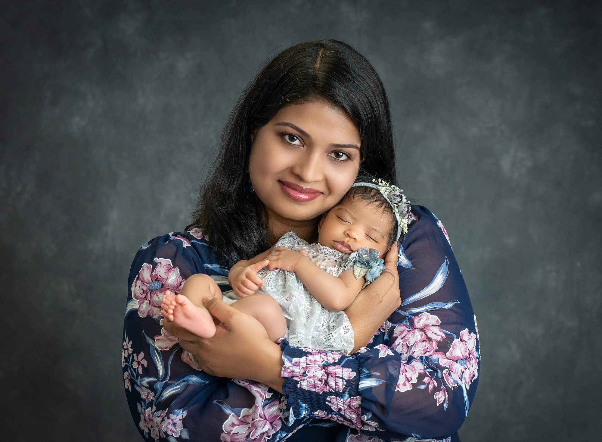 new mom cradling newborn baby girl in grey floral dress close to her chest