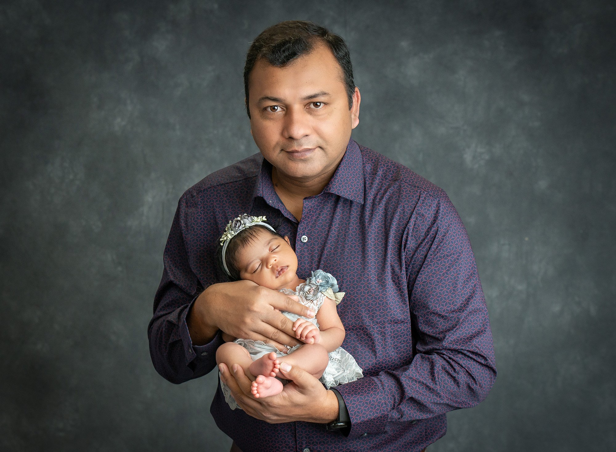 new dad holding sweet newborn baby girl dressed in a floral grey dress and headband on grey background