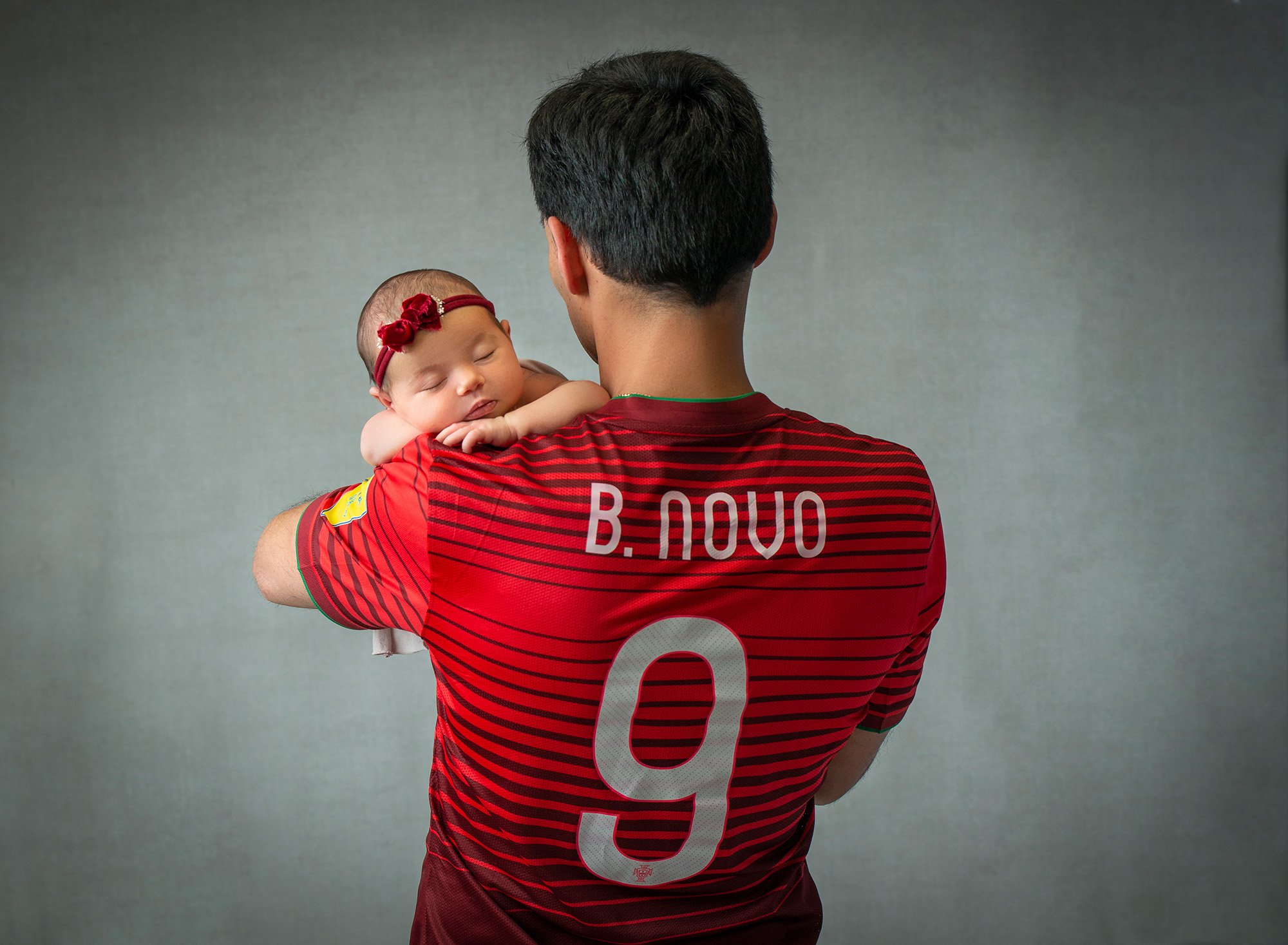 newborn baby girl wearing red bow asleep in dad's arms while he shows off his favorite sports jersey