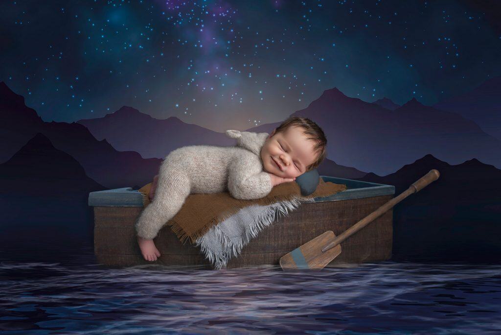 newborn baby boy sound asleep in a sweater romper on top of a row boat on an ocean with a starry mountain background