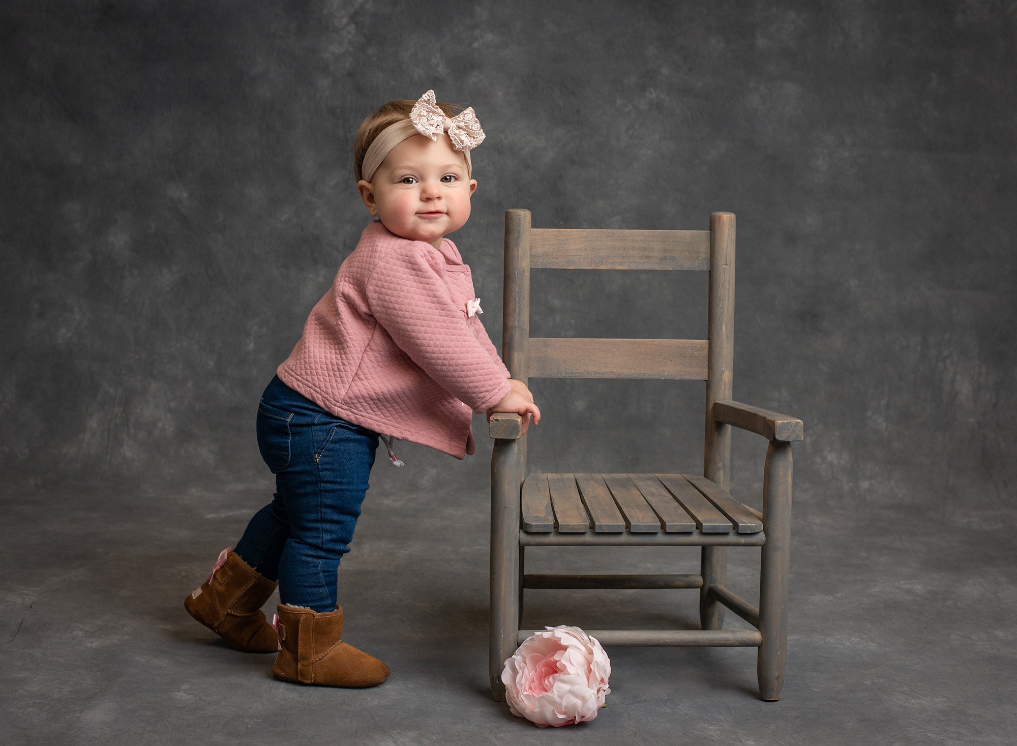 baby girl wearing pink button up and boots holding on to a rustic chair with pink flower in front on a dark gray backdrop