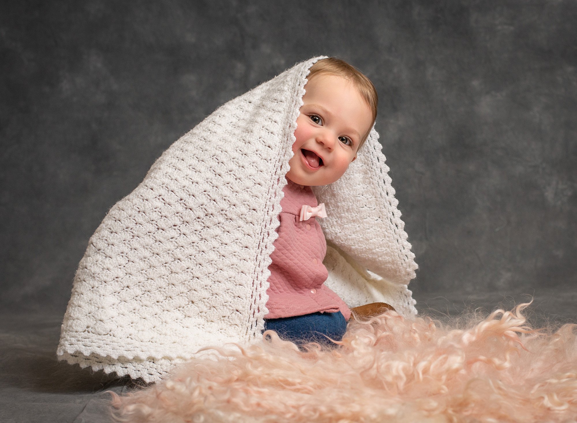 smiling baby girl with white blanket over her sitting in front of a pink fuzzy blanket