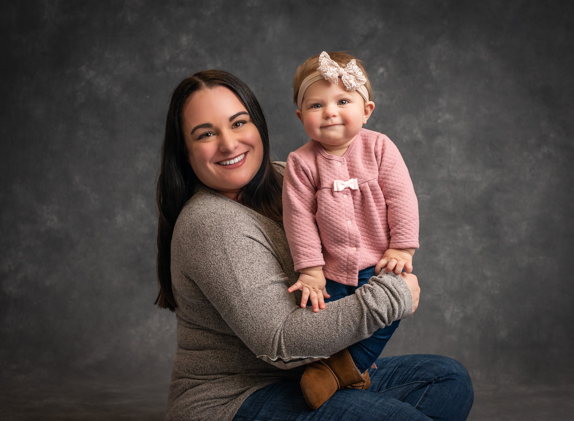smiling mom holding daughter wearing pink button up with dark gray backdrop