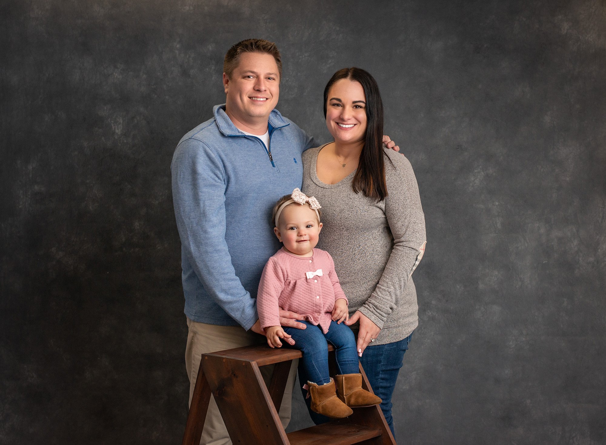 couple posing with their daughter who is sitting on a ladder with a dark gray backdrop