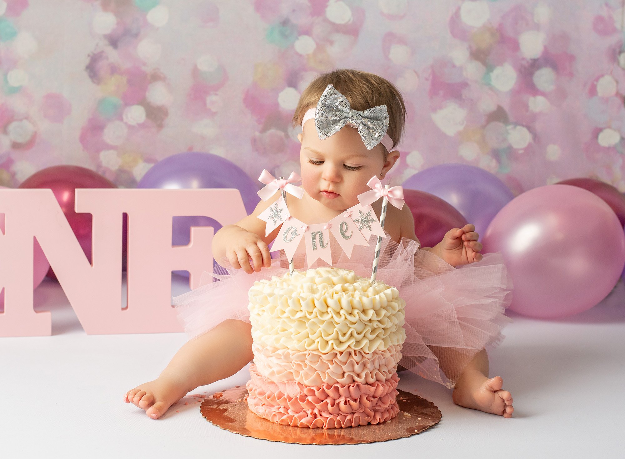 one year old baby girl wearing silver sequined bow and pink tutu surrounded by balloons and an ombre cake