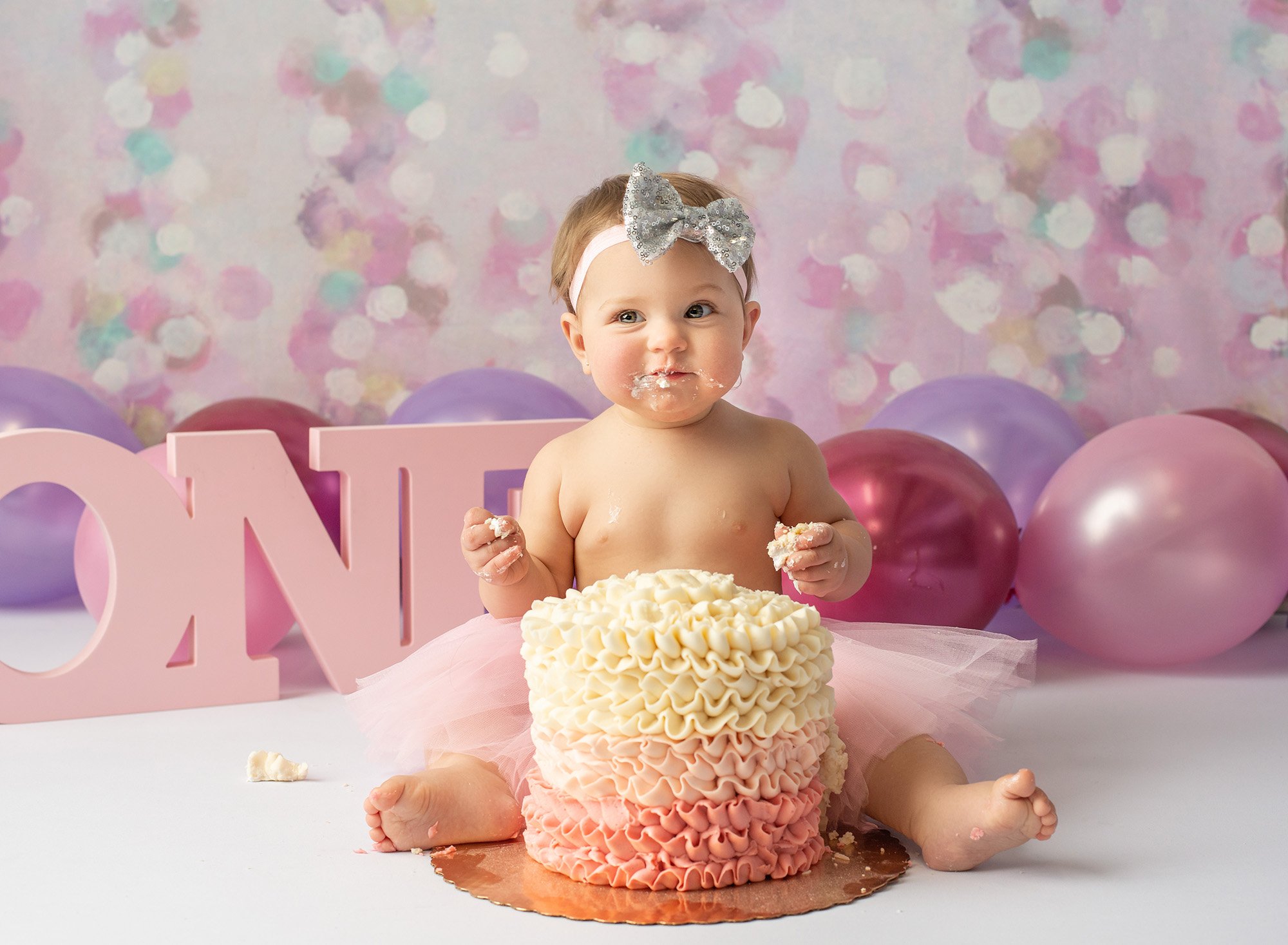 one year old girl with silver sequined bow, purple and pink balloons with ombre pink cake