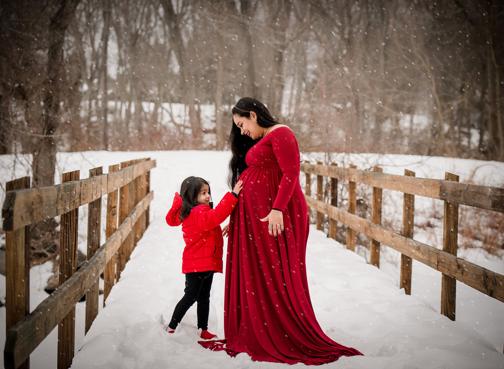 pregnant mom in red maternity gown on a snowy bridge surrounded by falling snow as daughter touches stomach
