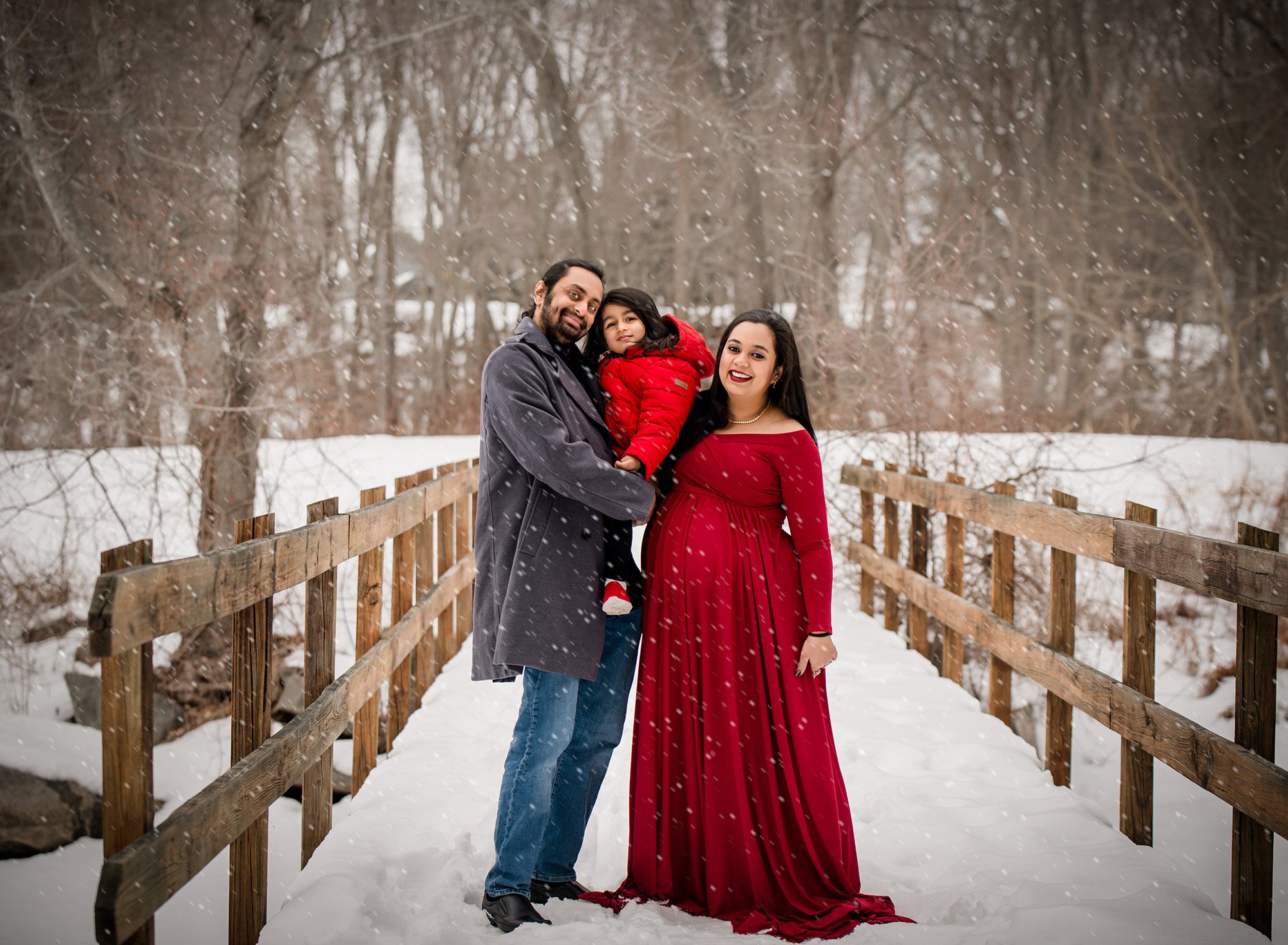 couples maternity photo including first child with red maternity dress on a snowy bridge with falling snow