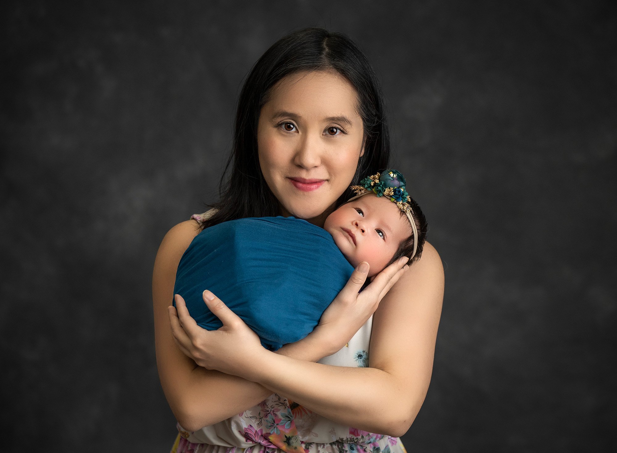 new mom cradling newborn baby girl swaddled in teal wearing teal floral headband on dark grey background