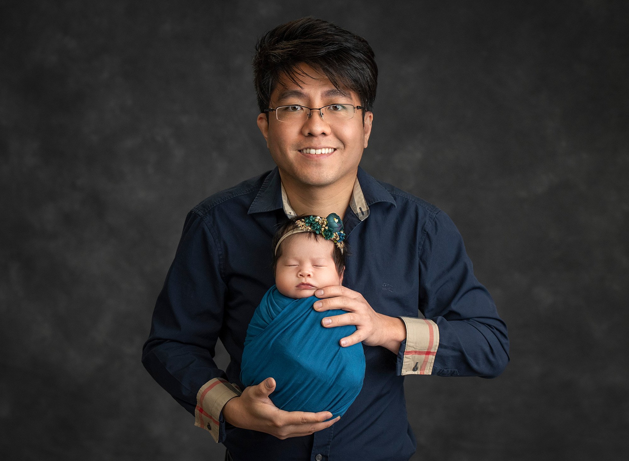 new dad wearing Burberry button up holding newborn baby girl swaddled in dark teal wrap wearing teal floral headband on dark grey background