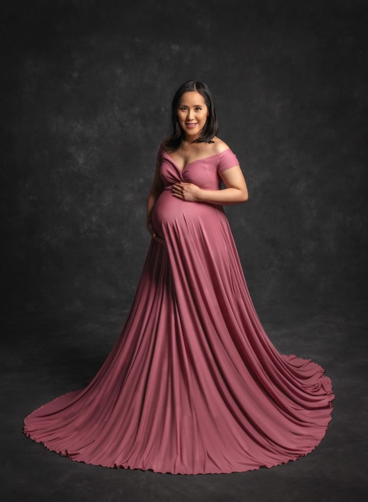 brunette pregnant woman wearing mauve flowing maternity dress on dark grey background