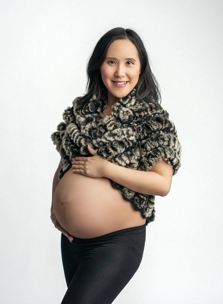 pregnant woman showing off stomach wearing black leggings and a faux fur sweater shirt on a white background