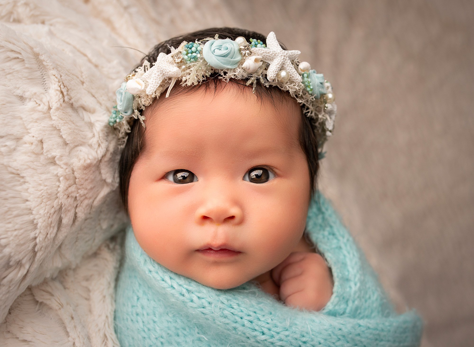 awake newborn baby girl swaddled in an aqua sweater wrap wearing starfish headband on a fluffy blanket