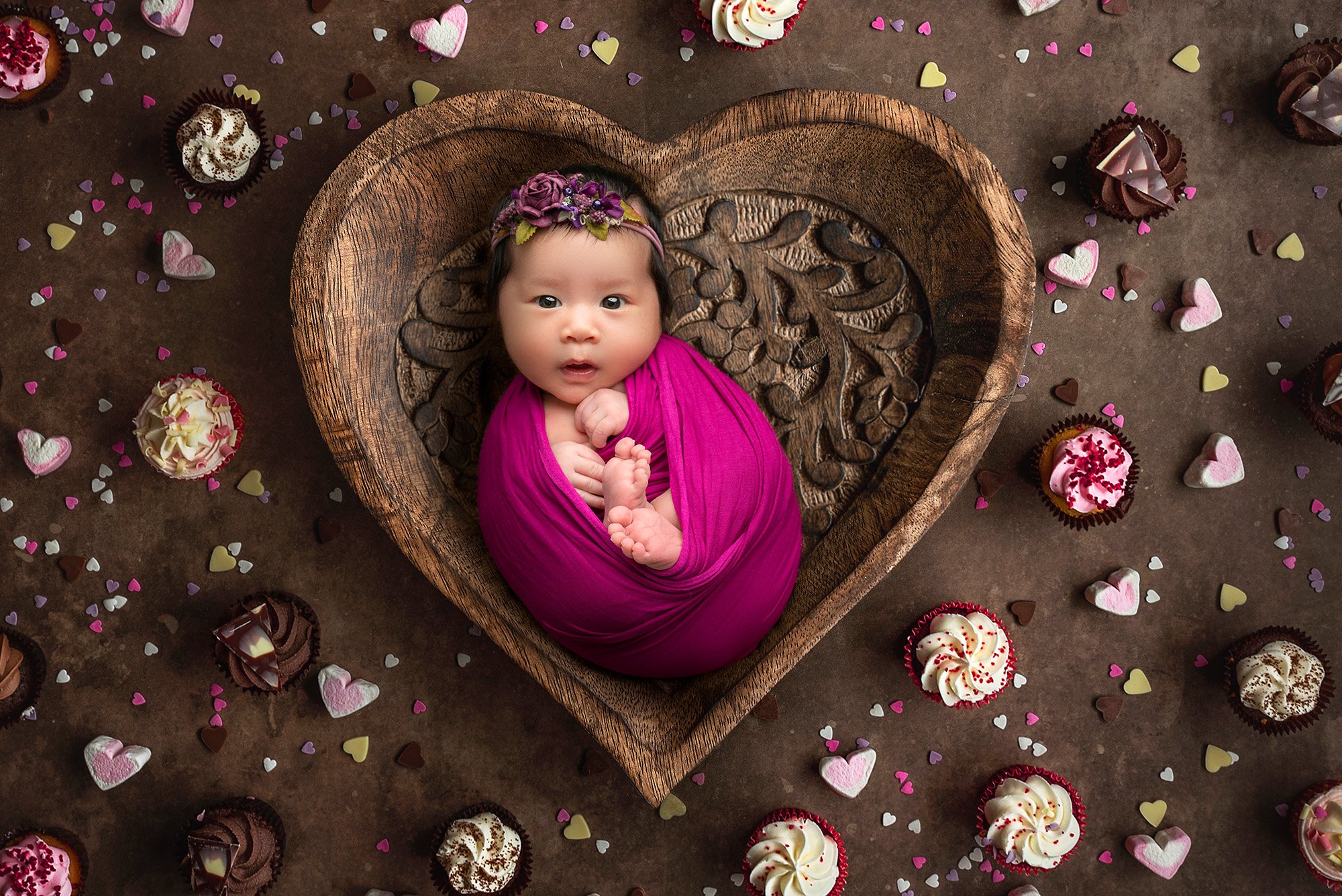 awake newborn baby girl swaddled in magenta wrap wearing purple floral headband laying in wooden heart bowl surrounded by cupcakes and heart candies