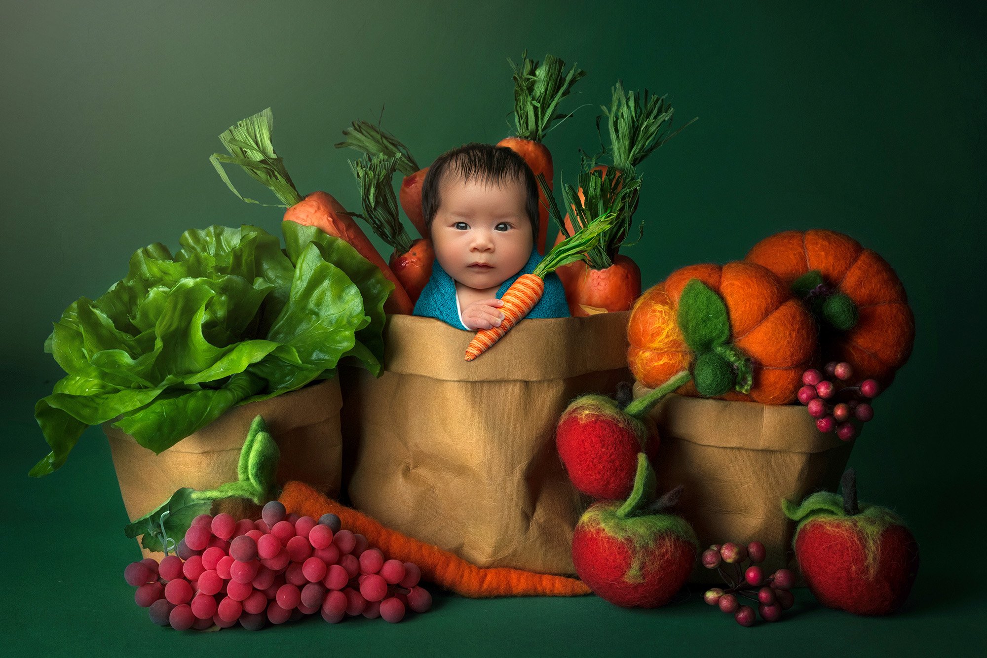 vibrant newborn photo newborn baby girl sitting in brown paper bag holding carrot surrounded by felt vegetables and fruit on a green background