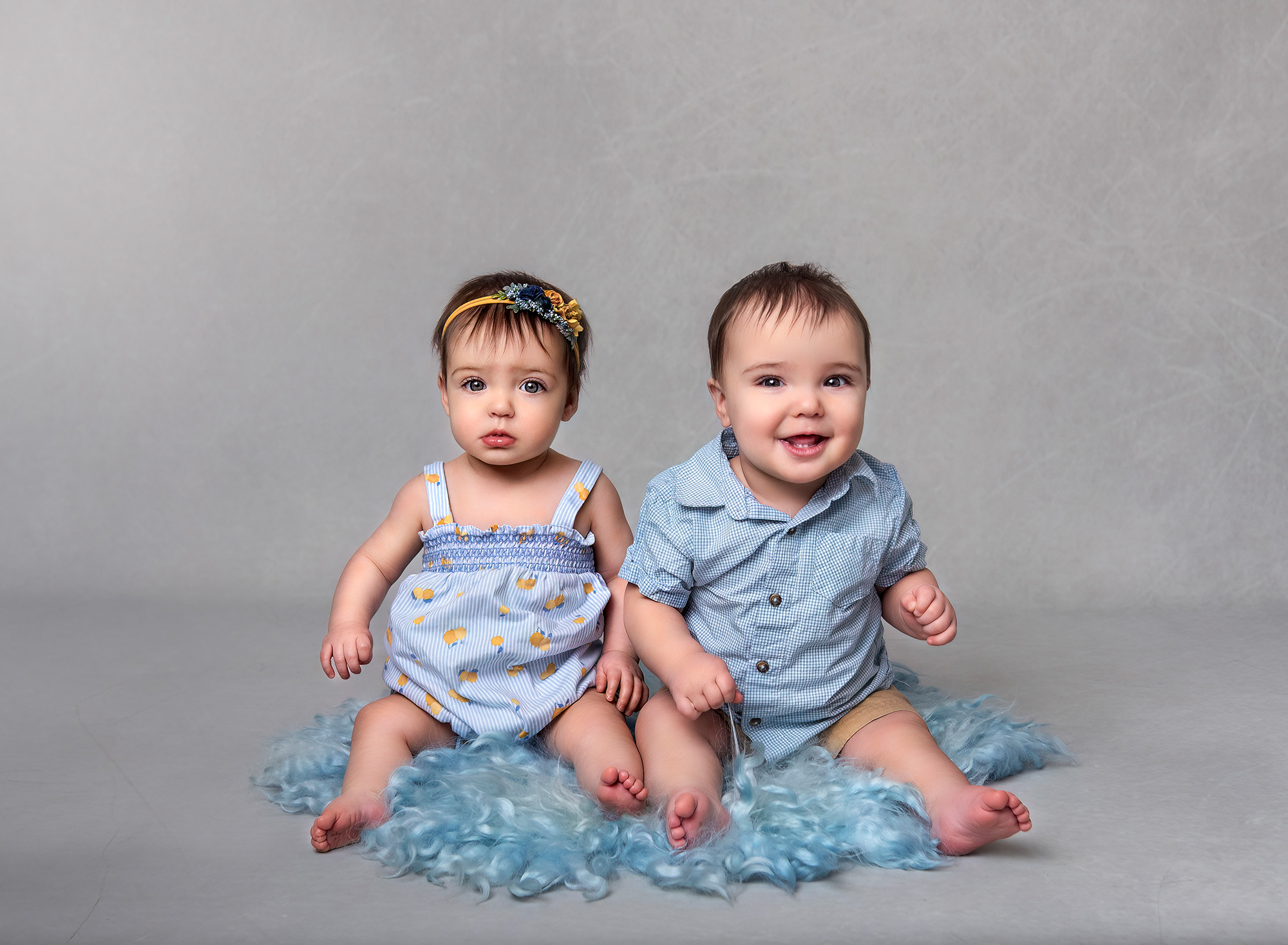 Twin boy and girl sitting on blue fur wearing blue outfits on a grey backdrop