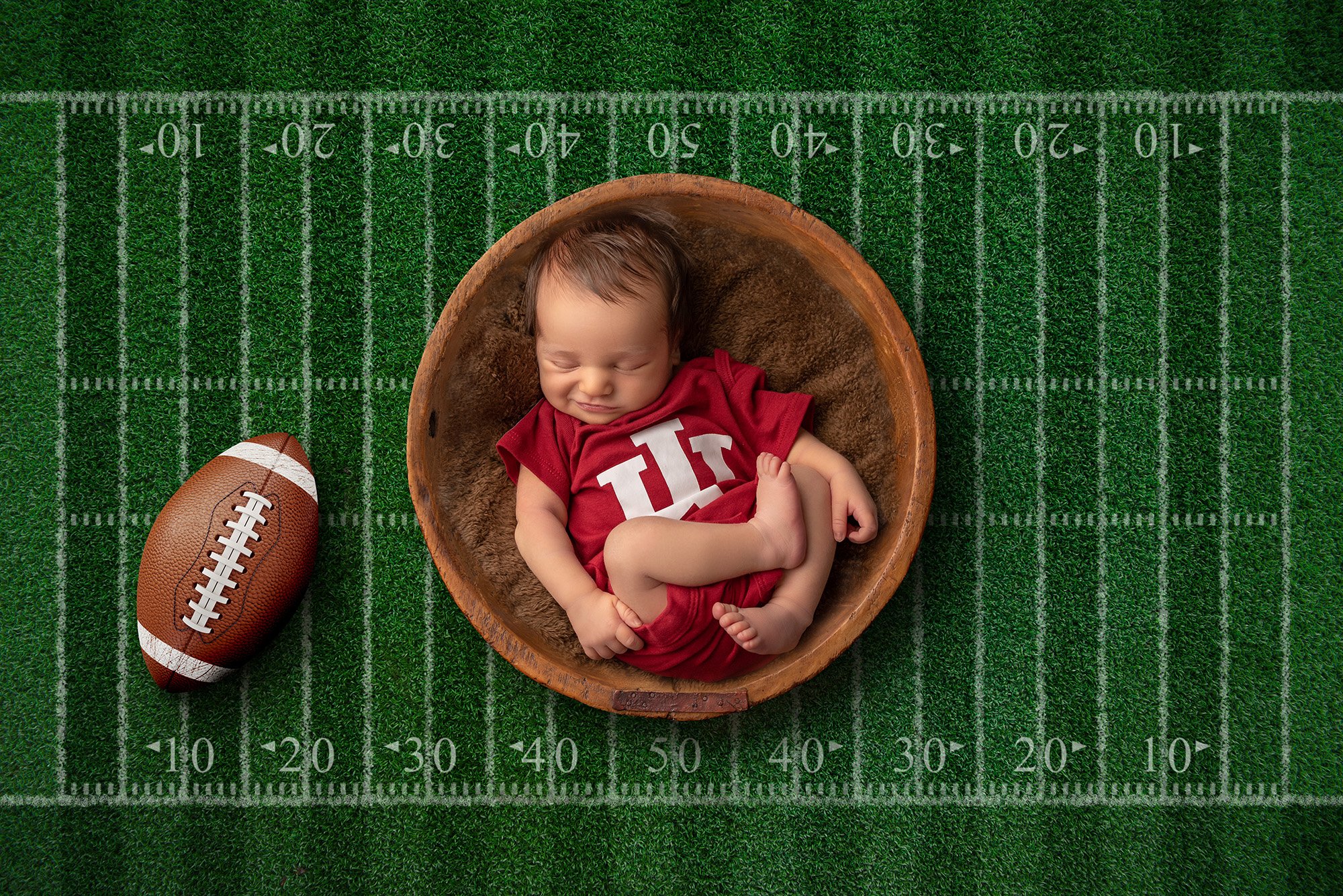 grinning newborn boy wearing a football jersey lying on a football field with a football