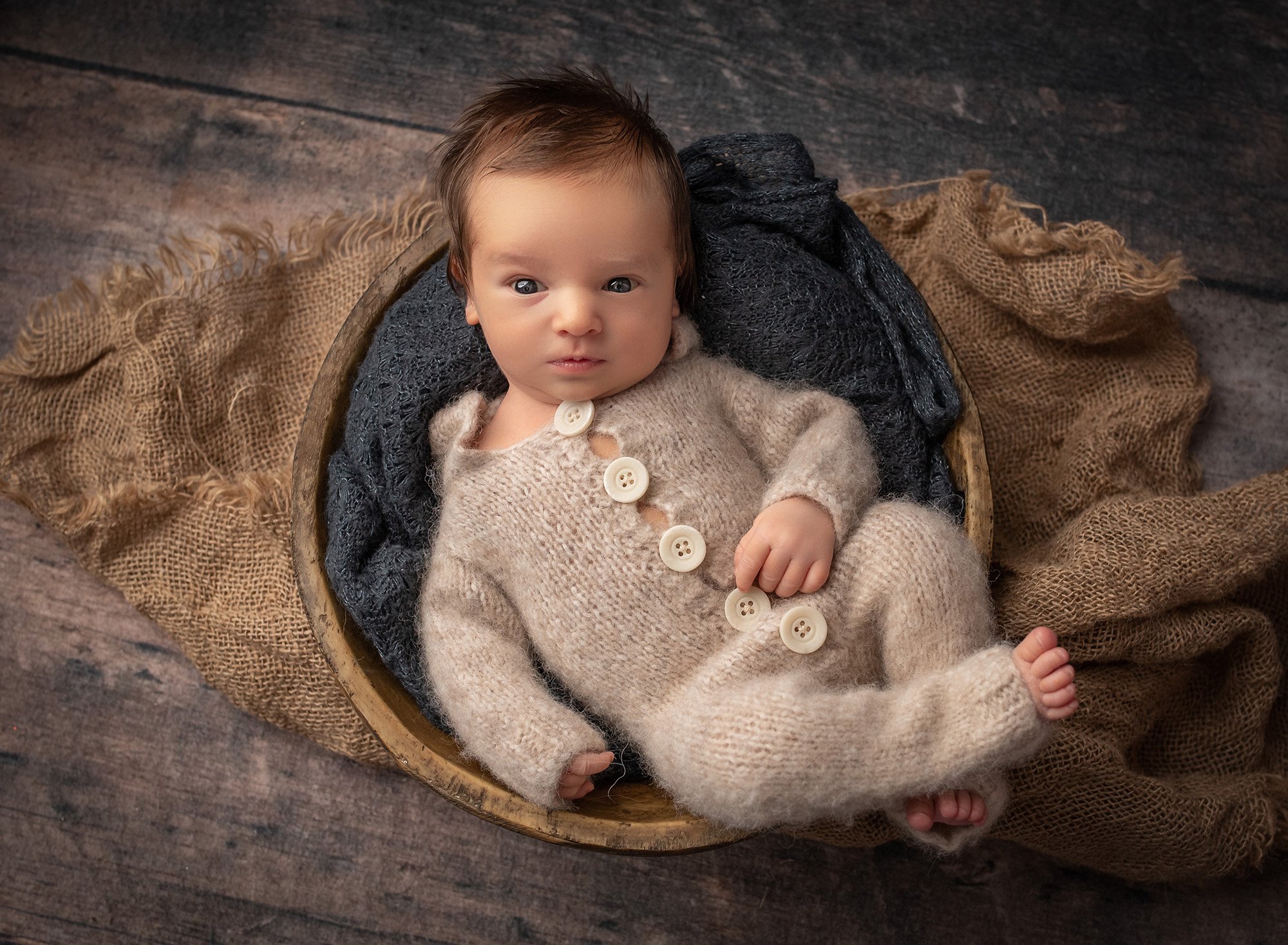 awake newborn baby boy lying in a wooden bowl wearing a knitted romper