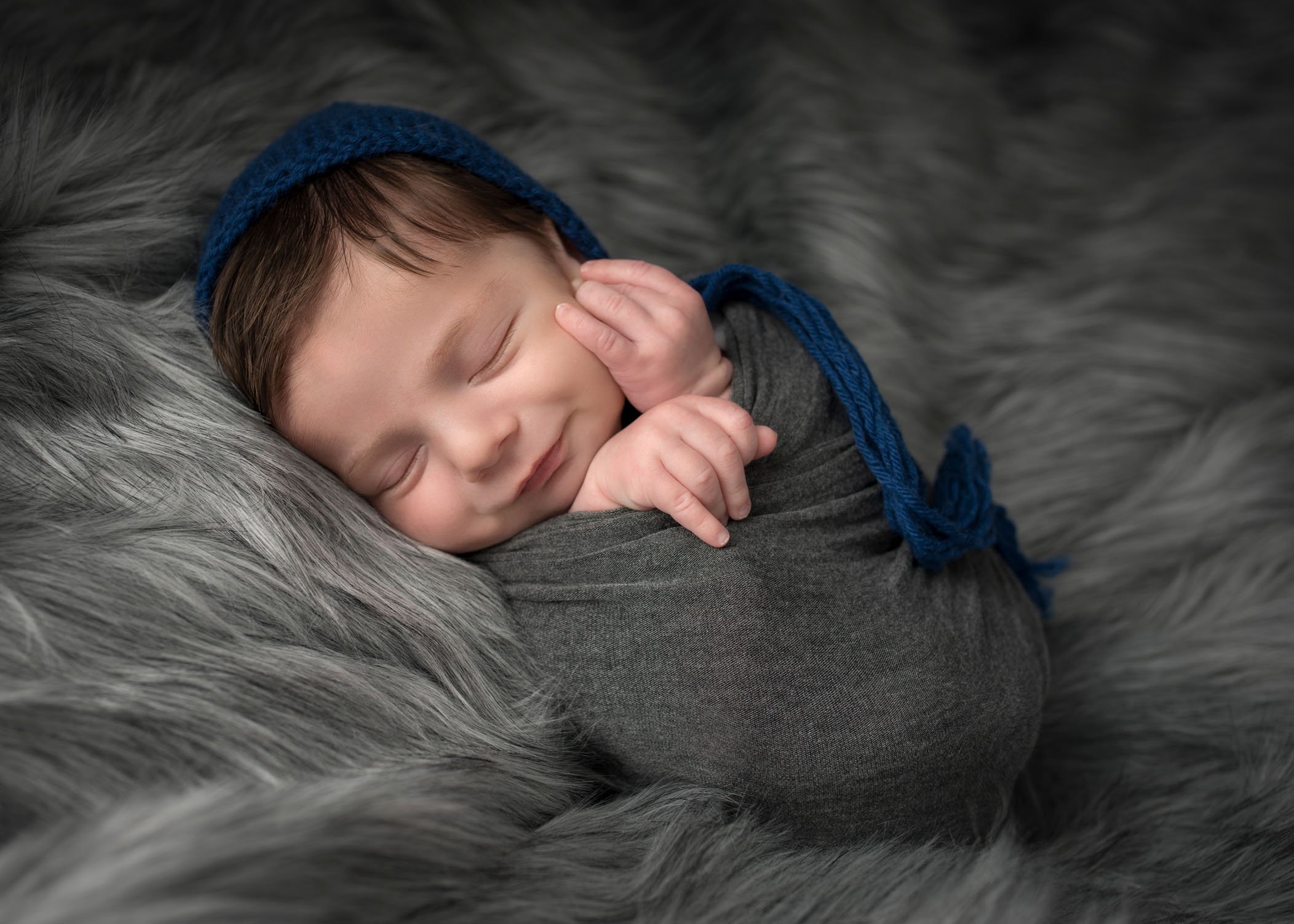 newborn boy swaddled, smiling while sleeping on grey fur