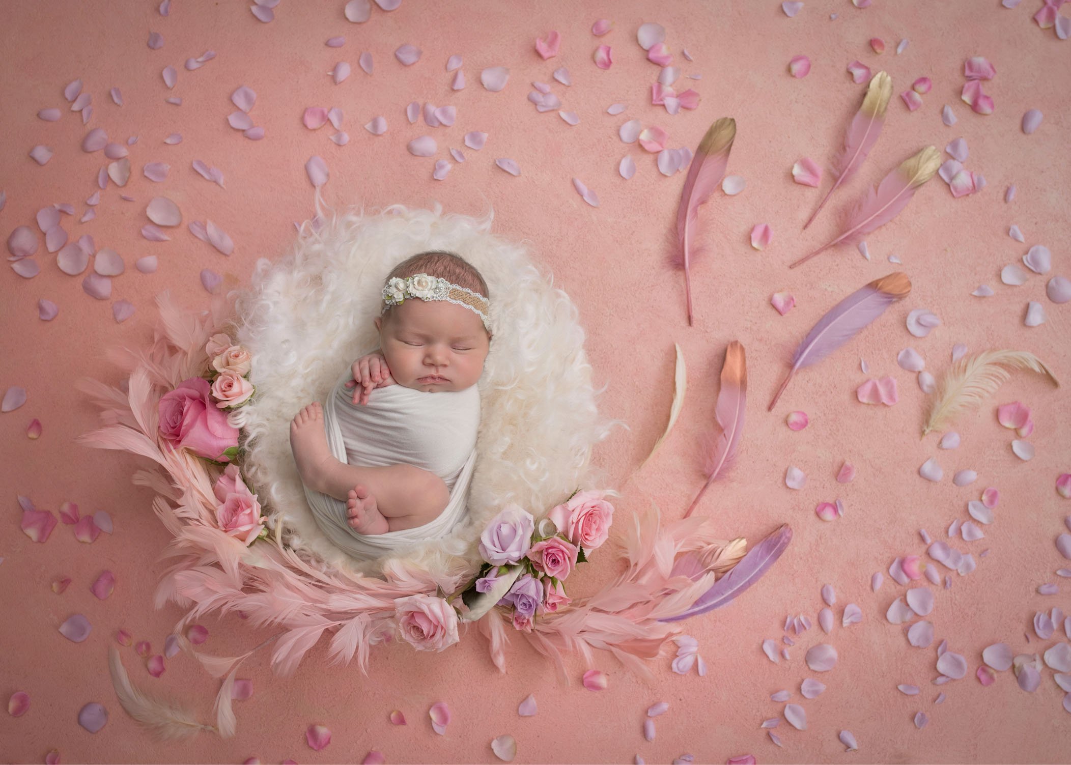 newborn baby girl sleeping in cream fluff on pink background with feathers and flowers