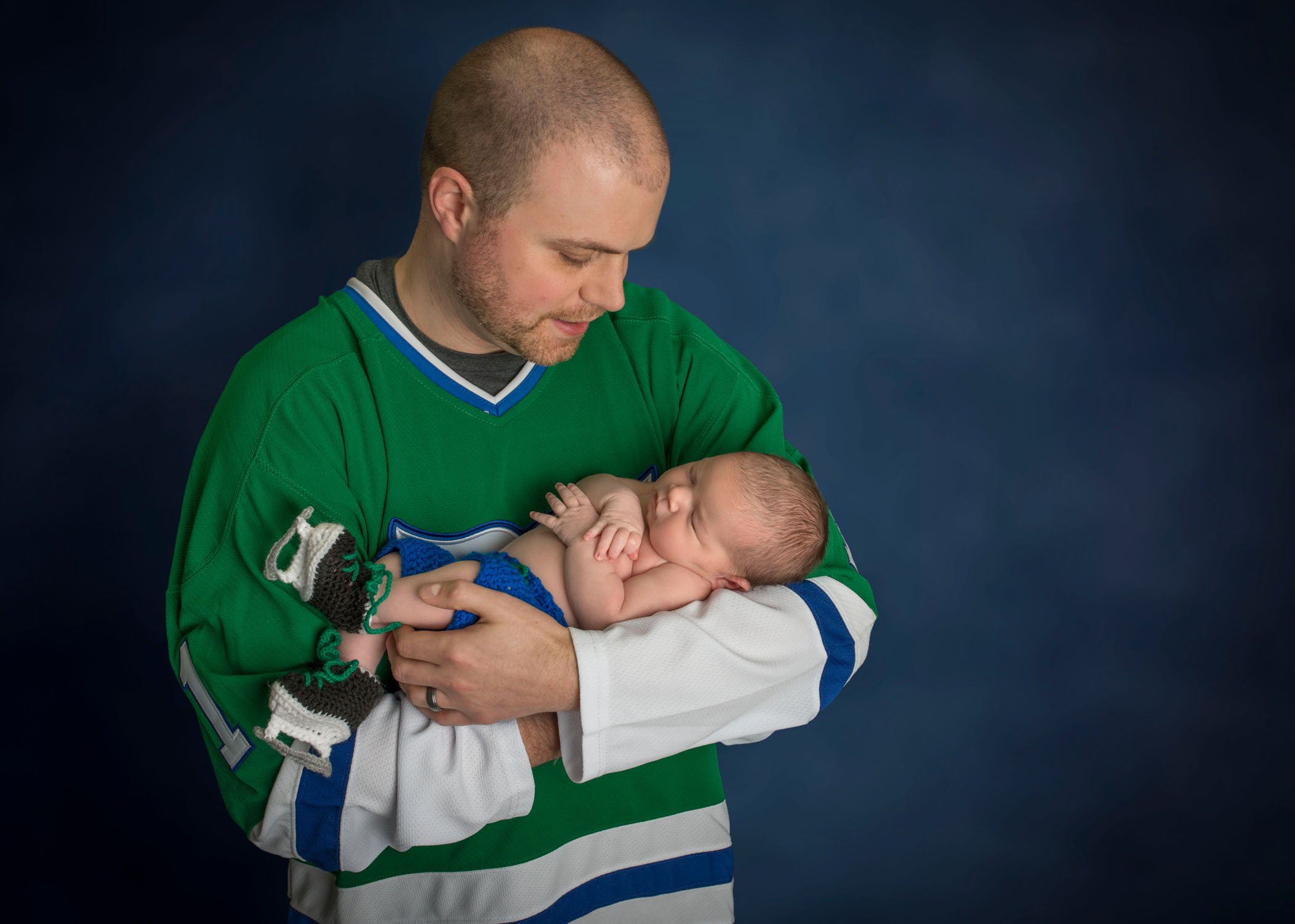 Dad holding newborn daughter while wearing ice hockey jersey and baby in crocheted ice skates
