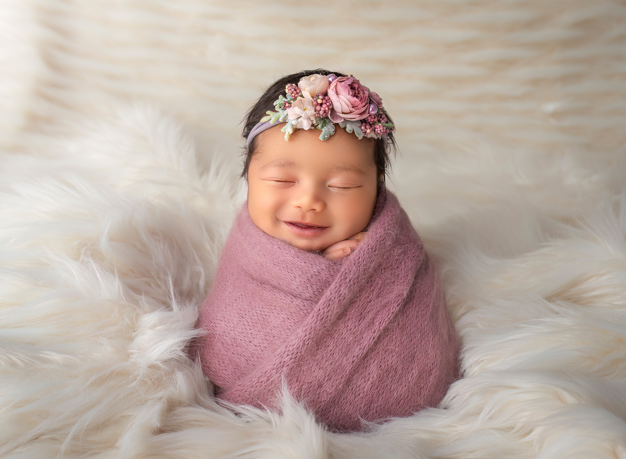 Newborn baby girl wrapped in purple, sitting on cream fur, smiling