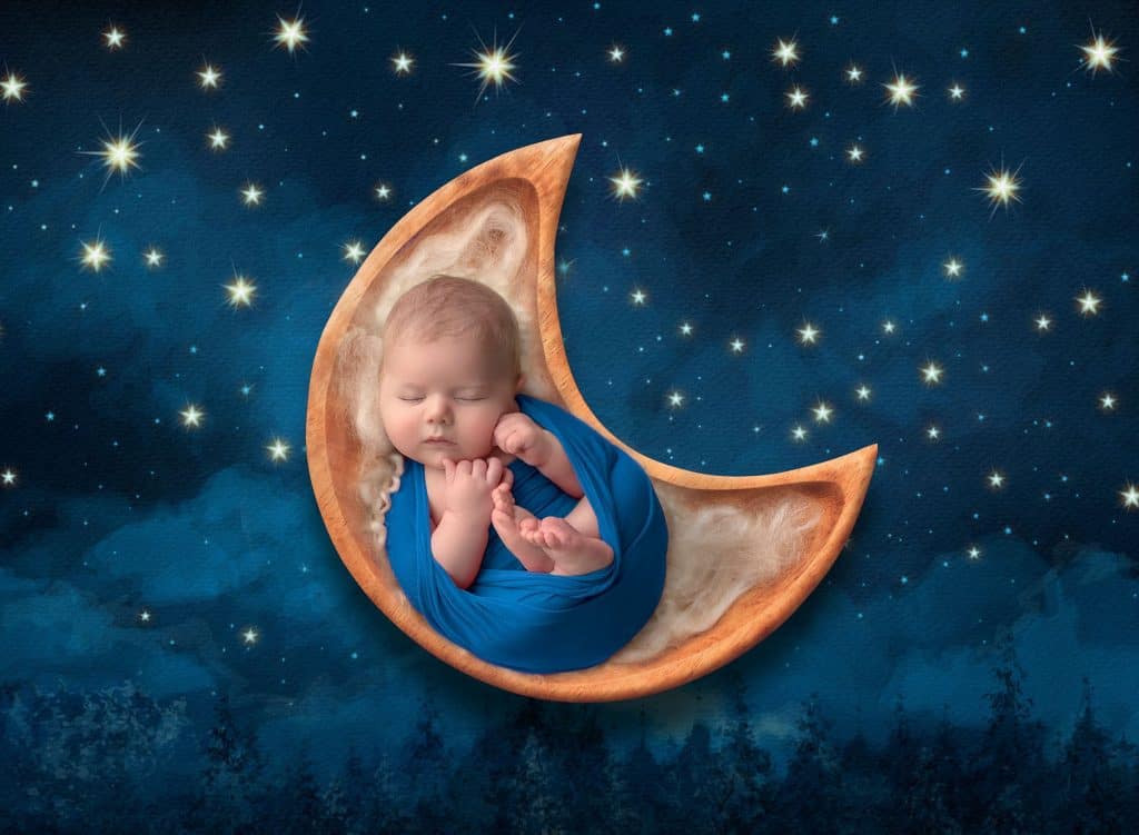 older newborn baby boy swaddled in deep blue sleeping in a moon prop on a night sky background best time to take newborn photos