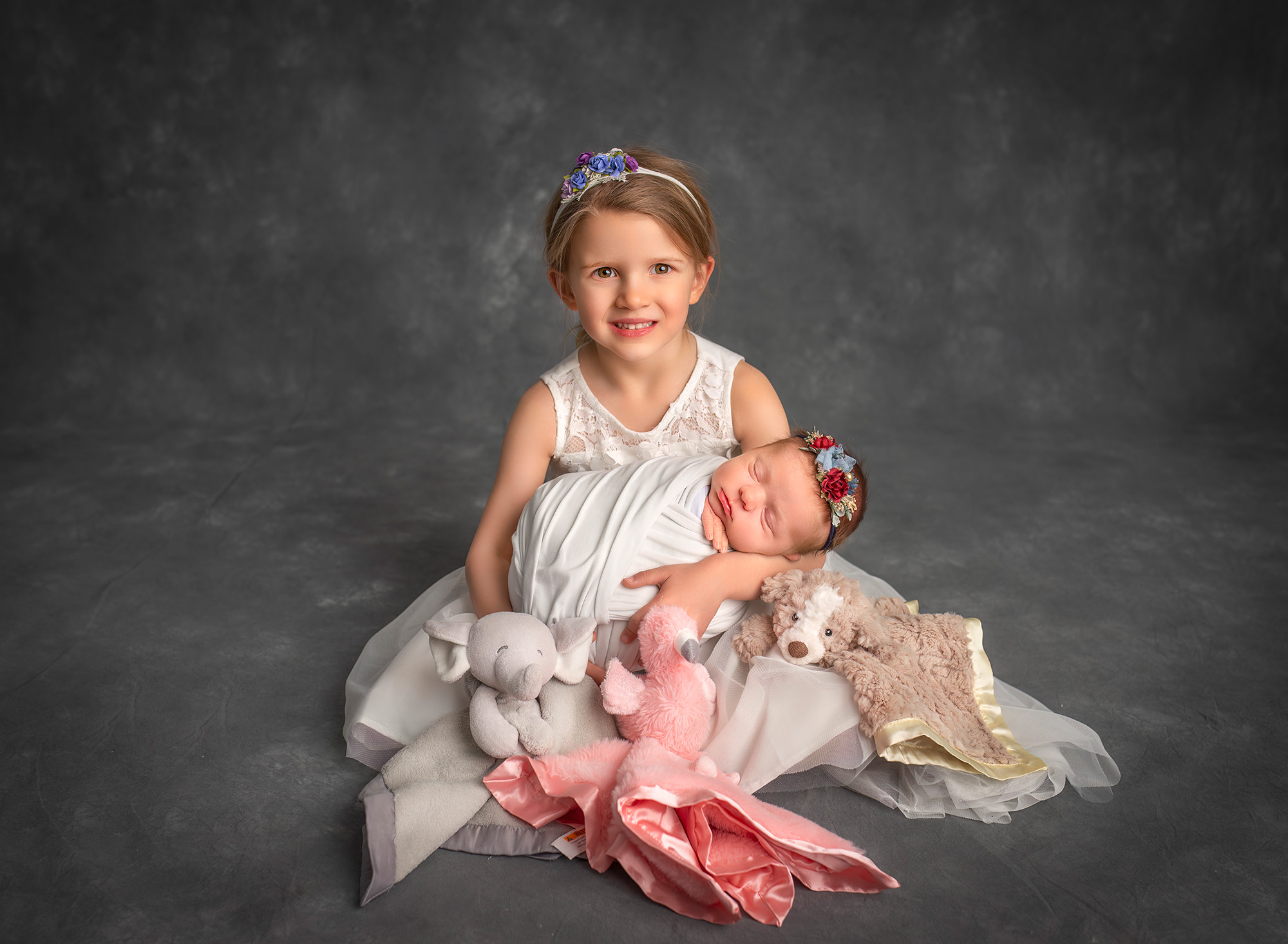 big sister and newborn big sister holding newborn sister in her arms with their stuffie toys next to them