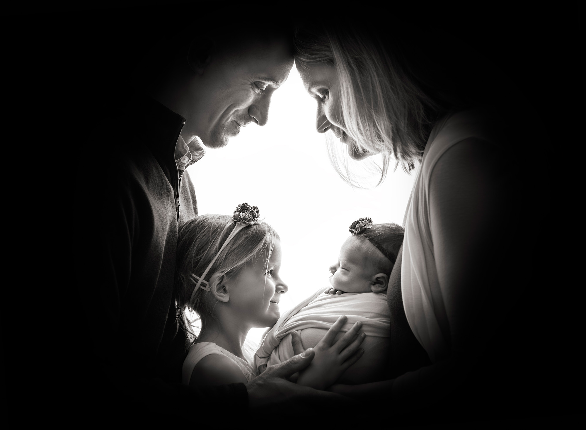 maternity and newborn photoshoot mom, dad and big sister and newborn baby all looking at the baby and smiling in black and white photograph