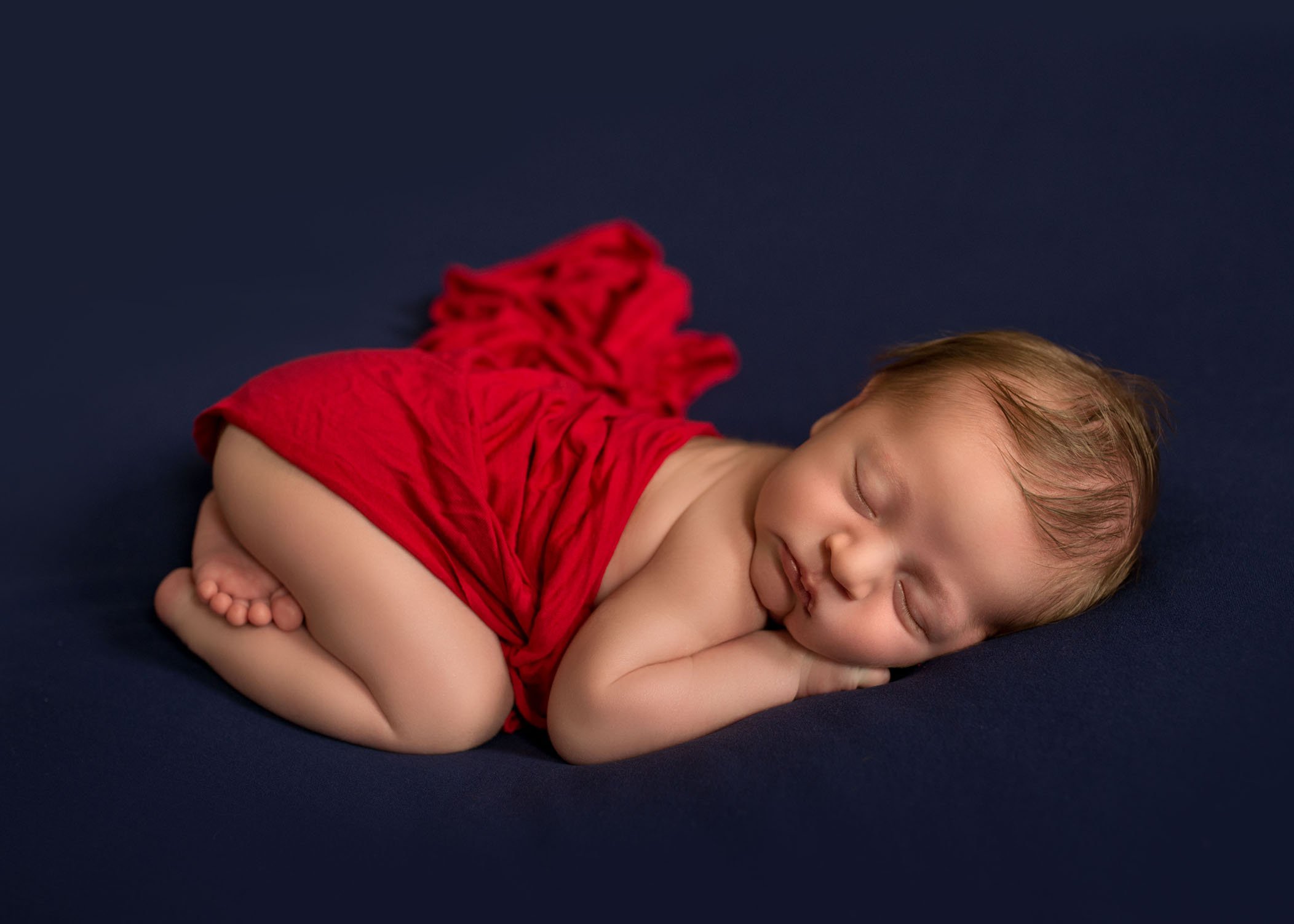 newborn baby boy sleeping on tummy on dark blue with red wrap