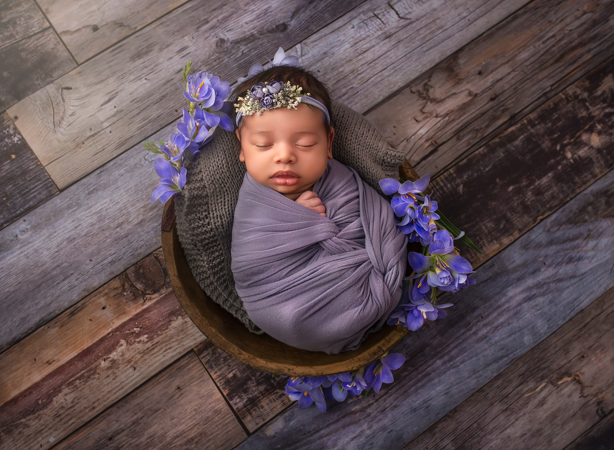 newborn photoshoot baby girl wrapped in purple swaddle sleeping in a wooden bowl on a wooden background