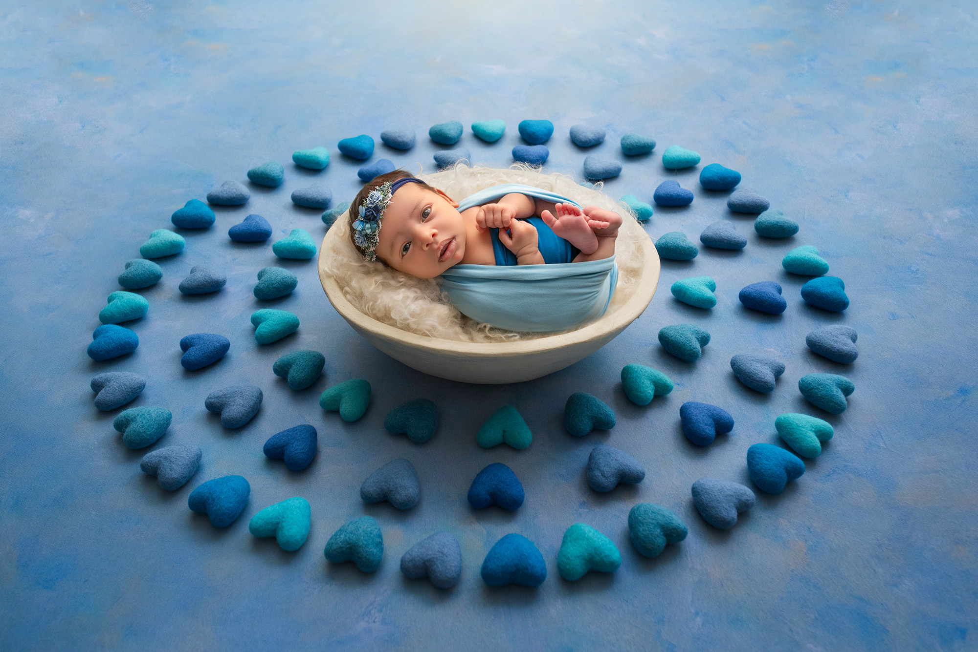 newborn photoshoot awake baby girl resting in a bowl surrounded by lots of blue felt hearts