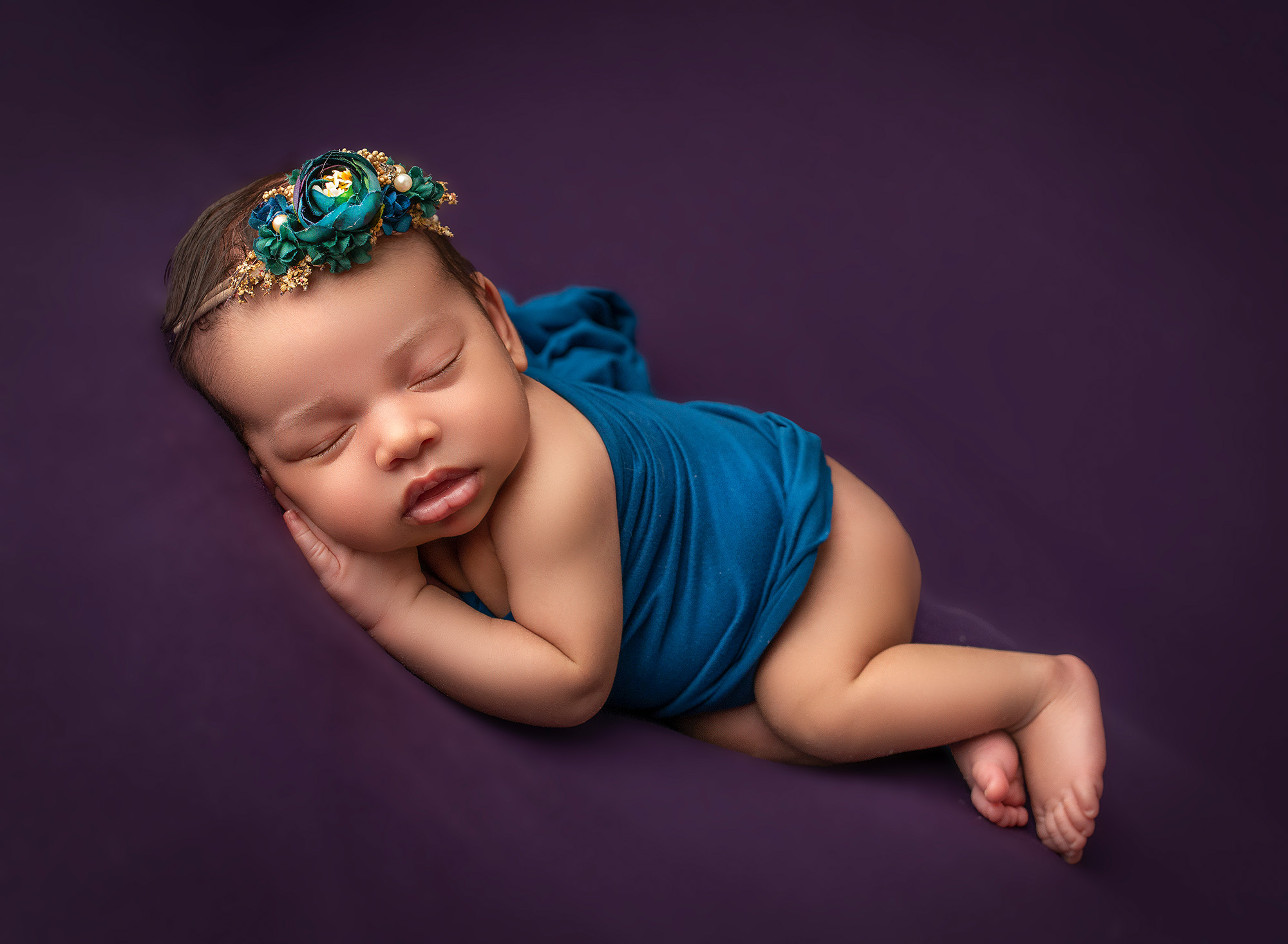 newborn photoshoot newborn baby girl asleep on a background of purple dressed in a teal wrap and floral headband