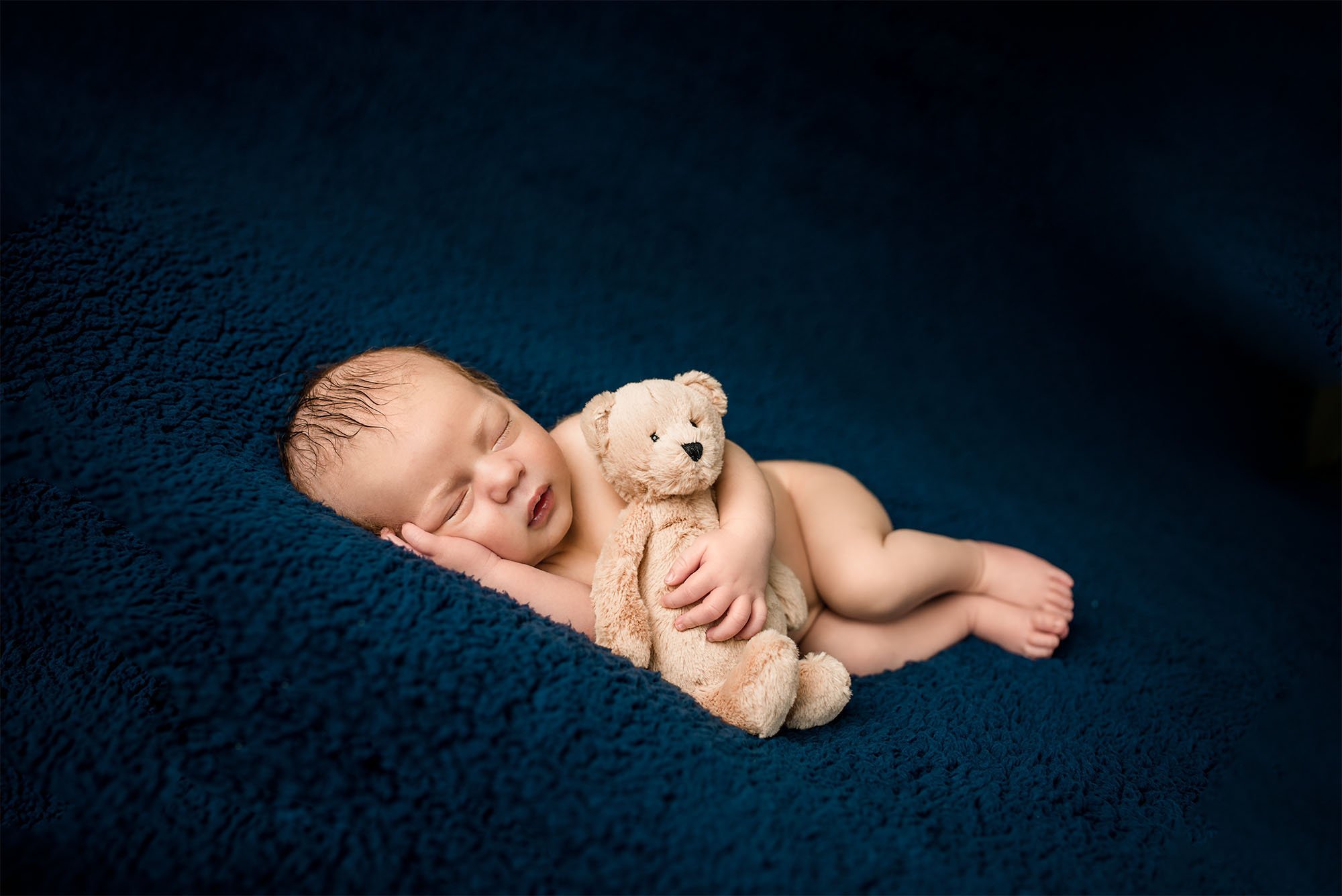 newborn baby sleeping on blue blanket with teddy under his arm