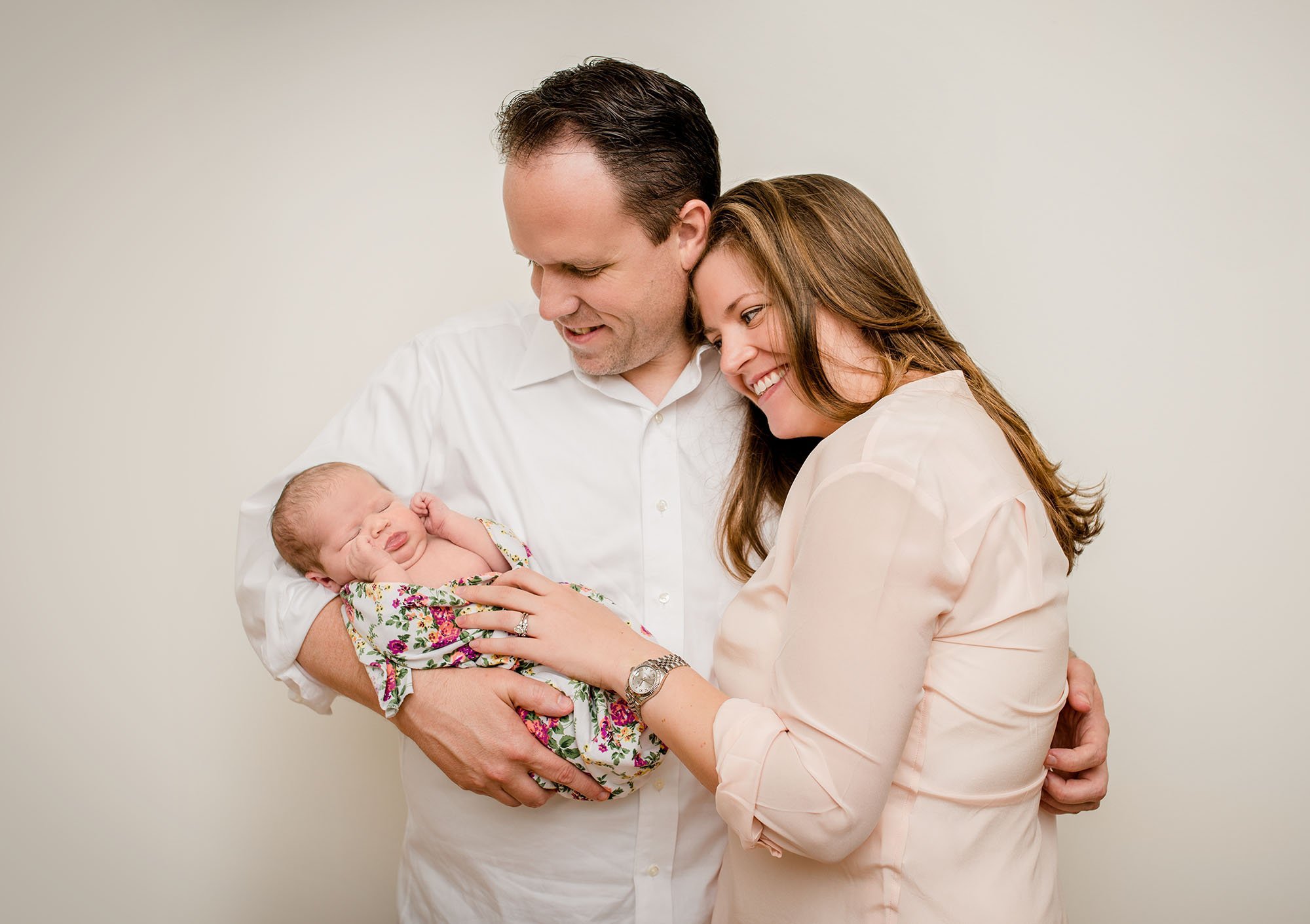parents holding newborn baby in arms smiling down at her One Big Happy Photo